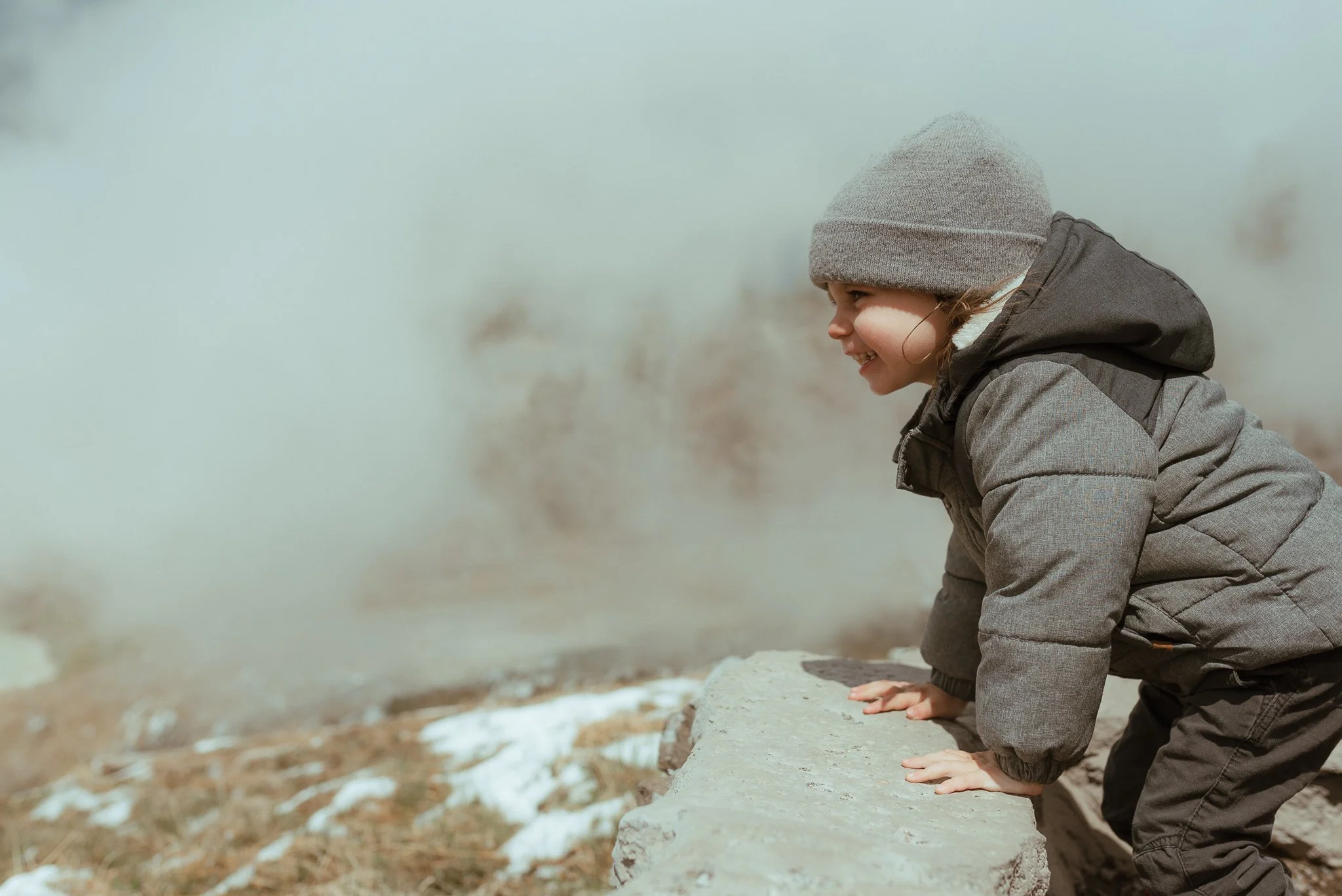 A young child wearing a gray knit cap and puffy jacket crouches on a rock along a riverbank, looking out at the water with a smile.
