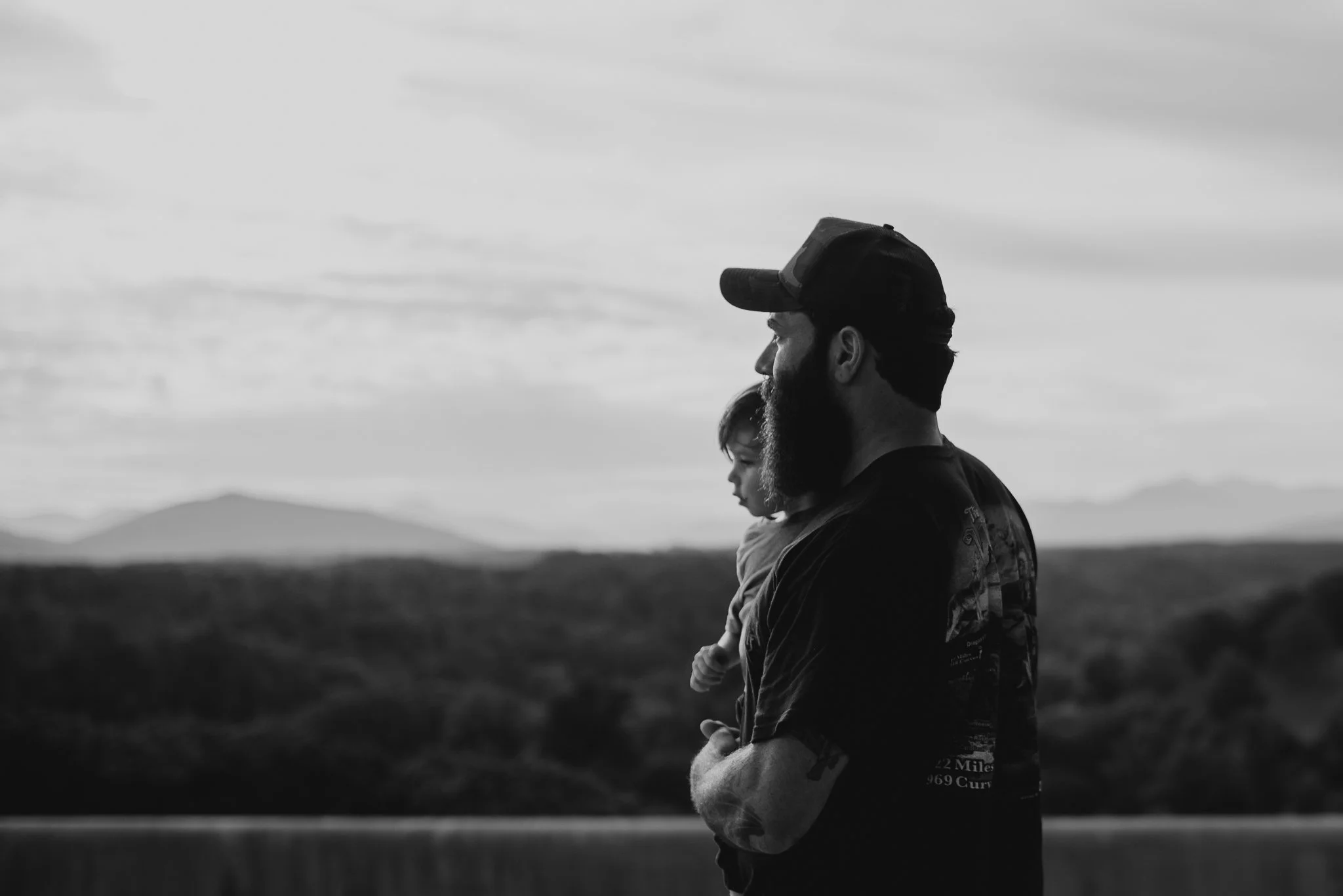 A bearded man and a young girl are standing outdoors in profile view, both looking to the right. The man is wearing a cap and has tattoos on his arm. The background shows open landscape with mountains in the distance and a cloudy sky.