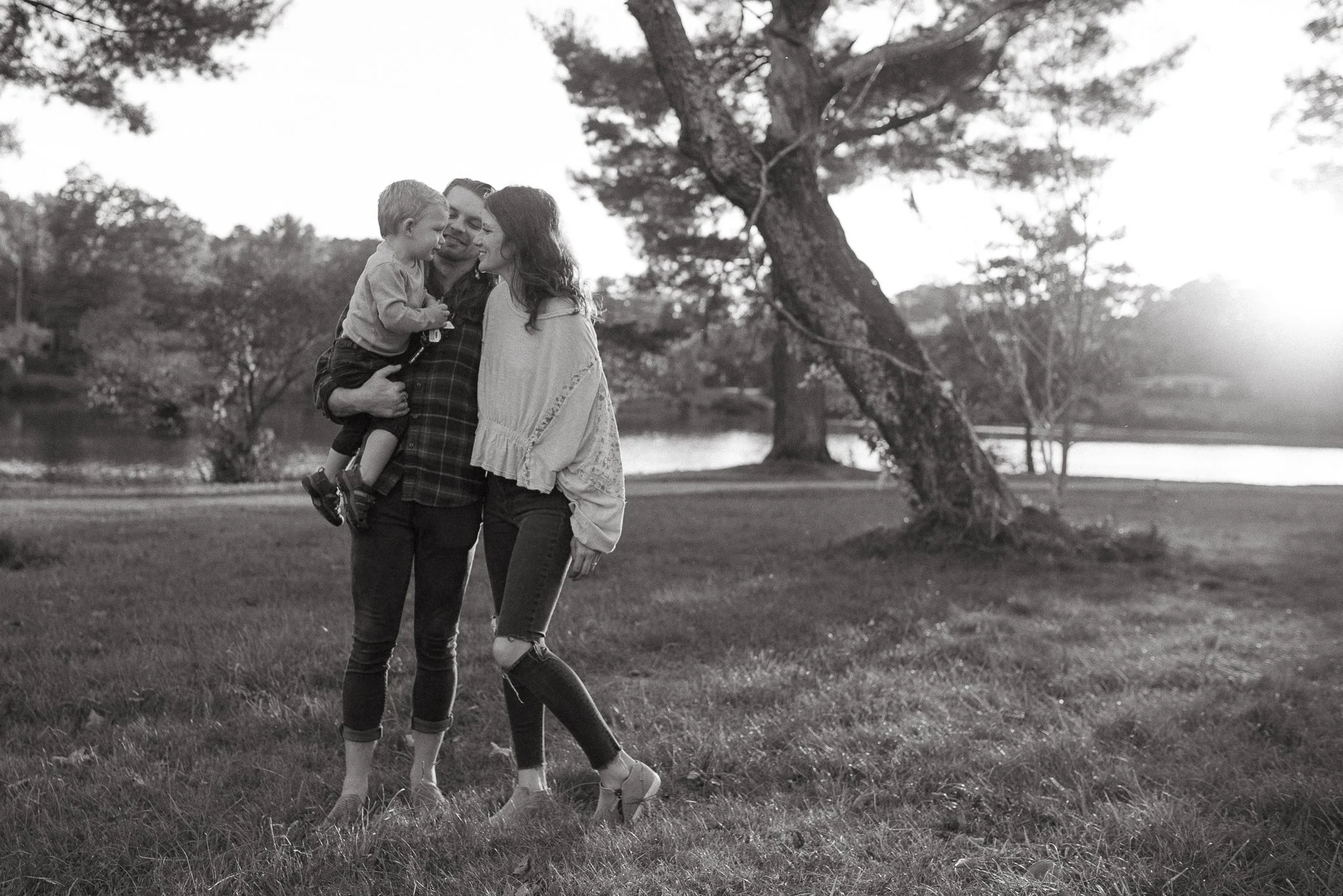 A black-and-white photo of a family standing outdoors on grass. The family consists of a man, woman, and a young boy, who are close together, smiling, and interacting affectionately. The family appears to be enjoying a moment in a park or natural set