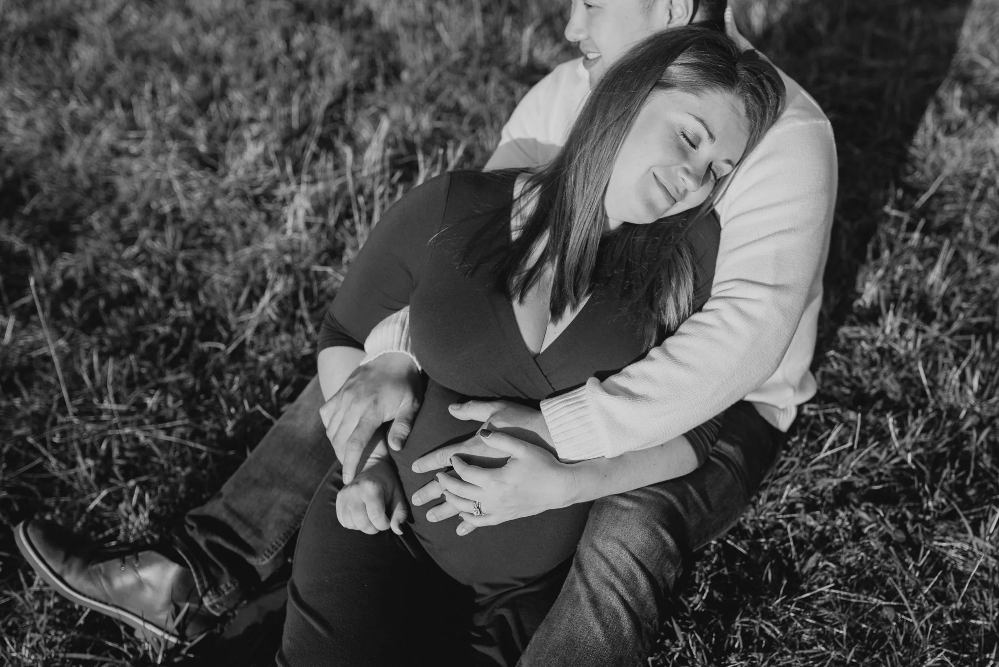 A black and white photo of a couple sitting on grass, with the woman resting her head on the man's chest, both smiling softly and appearing relaxed and happy.