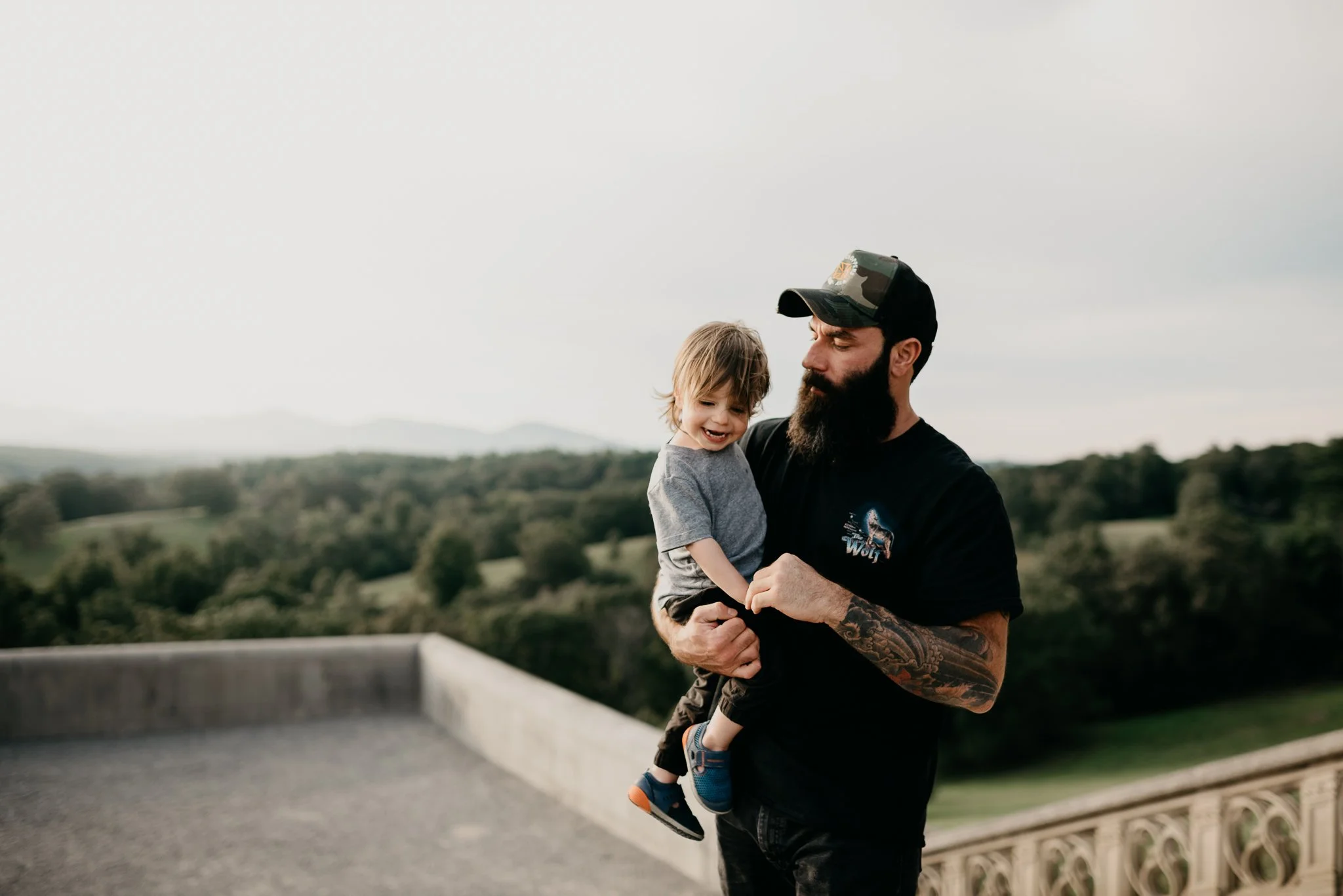 A bearded man with tattoos holding a smiling young boy with light brown hair and wearing a gray shirt outdoors on a cloudy day, with a scenic landscape of trees and hills in the background.