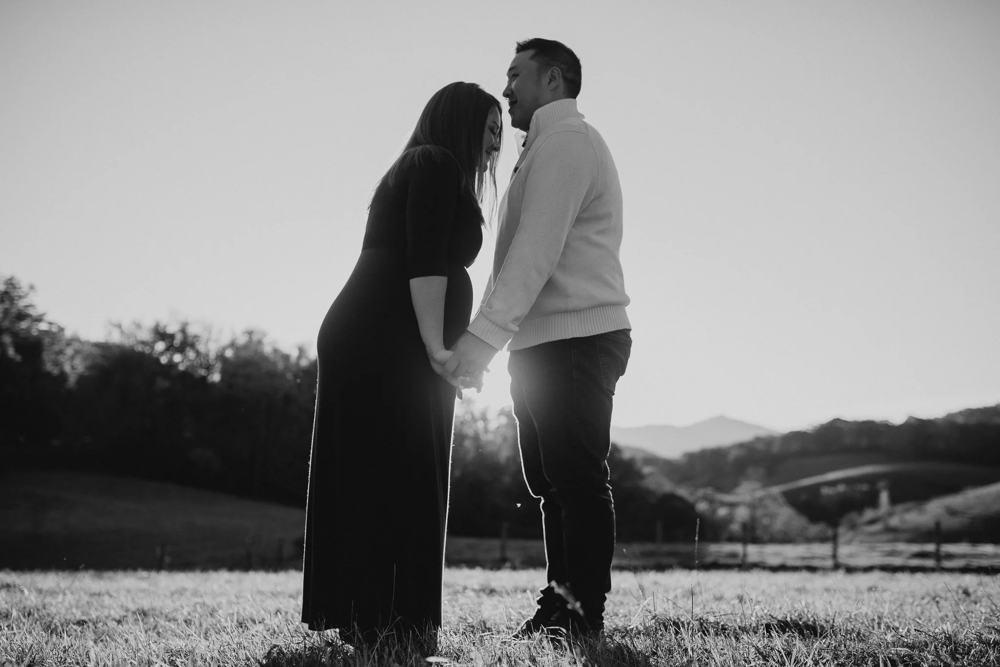 A black and white photo of a couple holding hands and leaning their foreheads together outdoors, with mountains and trees in the background.