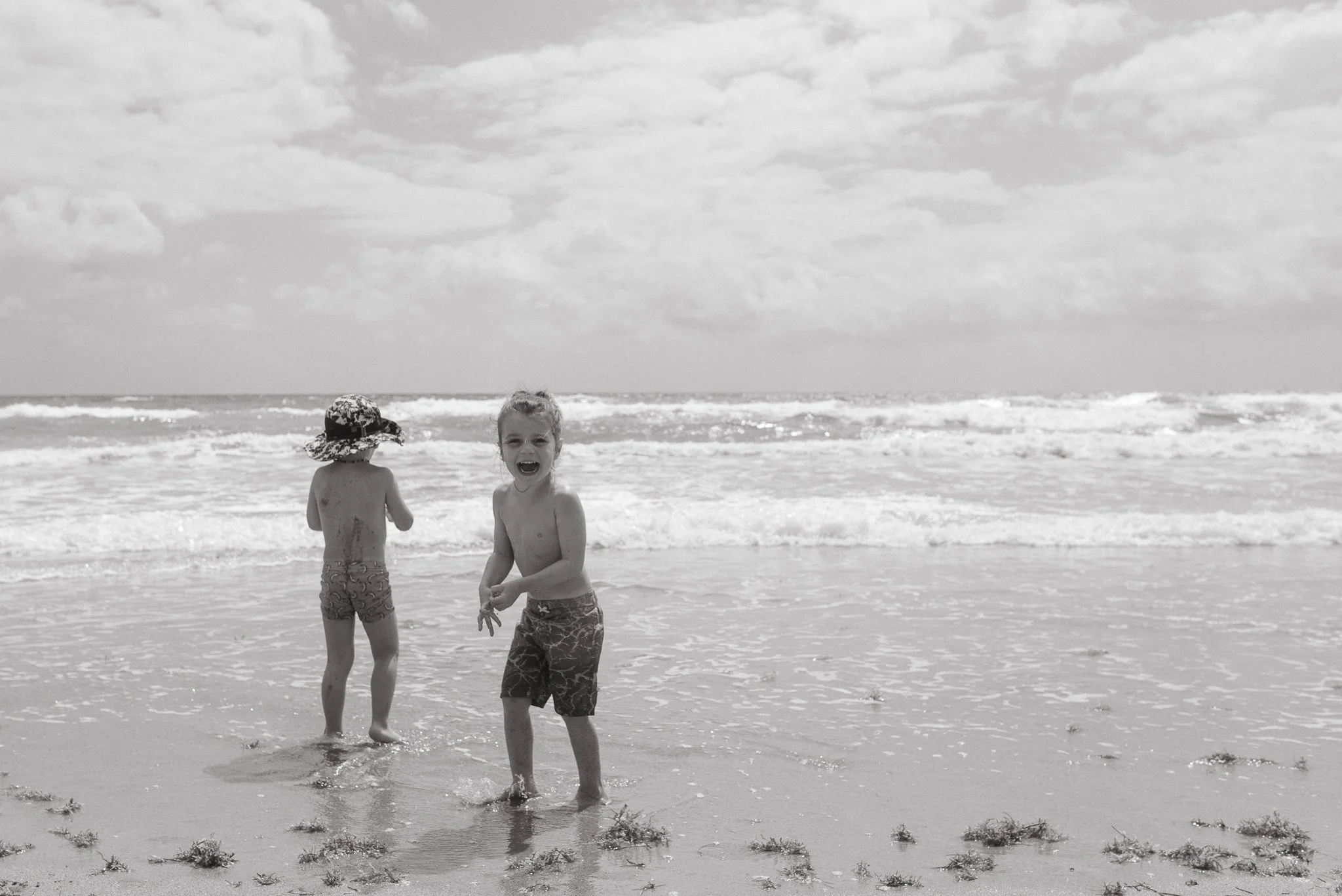 Two children playing in the ocean waves on the beach, one wearing a sun hat and the other smiling and barefoot in the water.