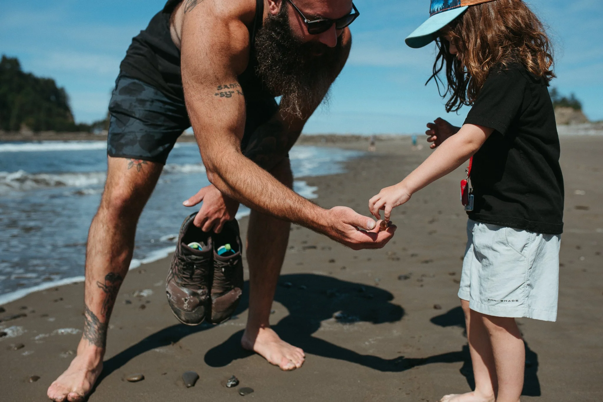 A man with a beard, sunglasses, and tattoos is crouched on the beach, holding the hand of a young girl with long hair and a cap, near the shoreline with rocks and trees in the background.