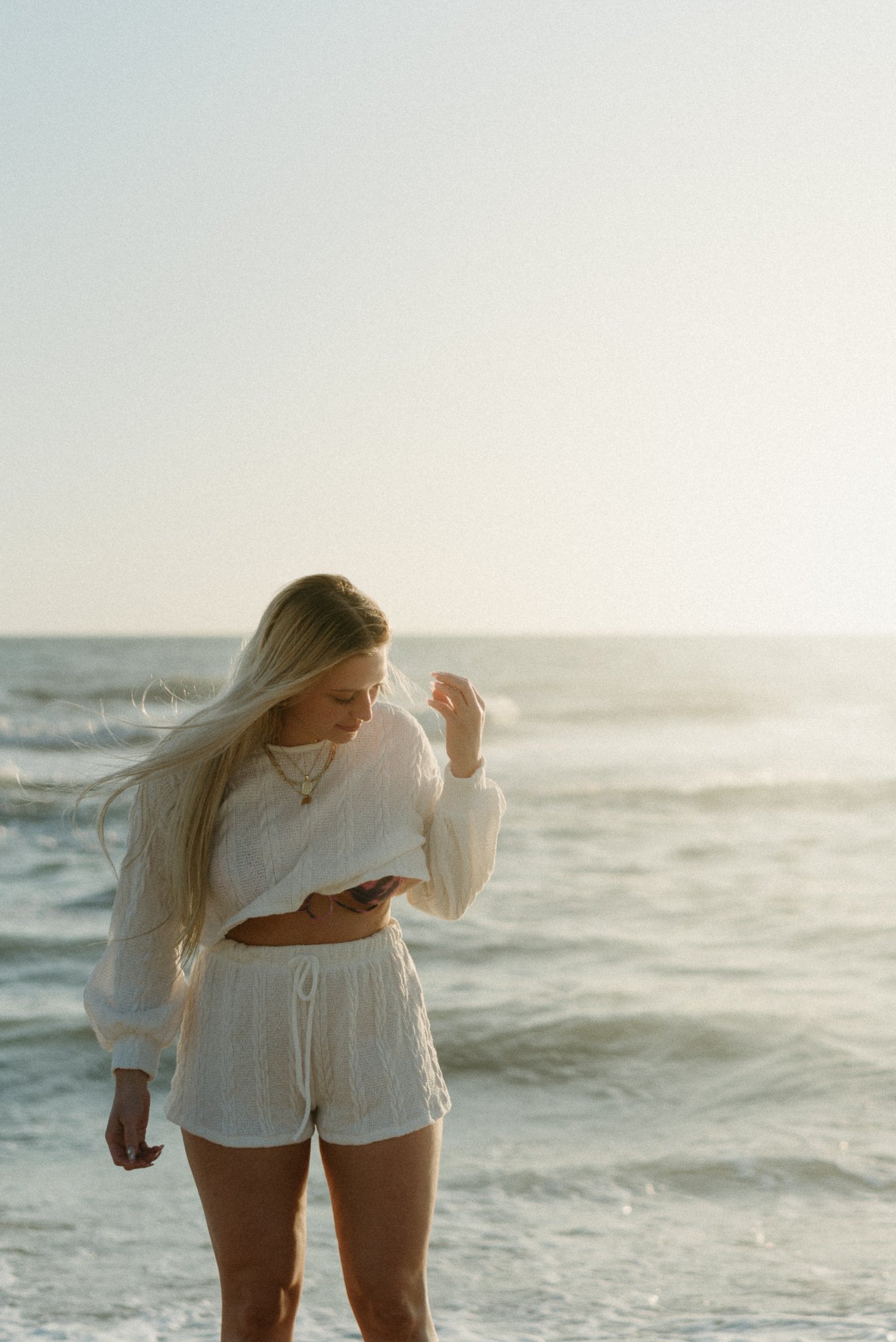 A woman with long blonde hair wearing a white sweater and shorts walking along the beach near the ocean during sunset.