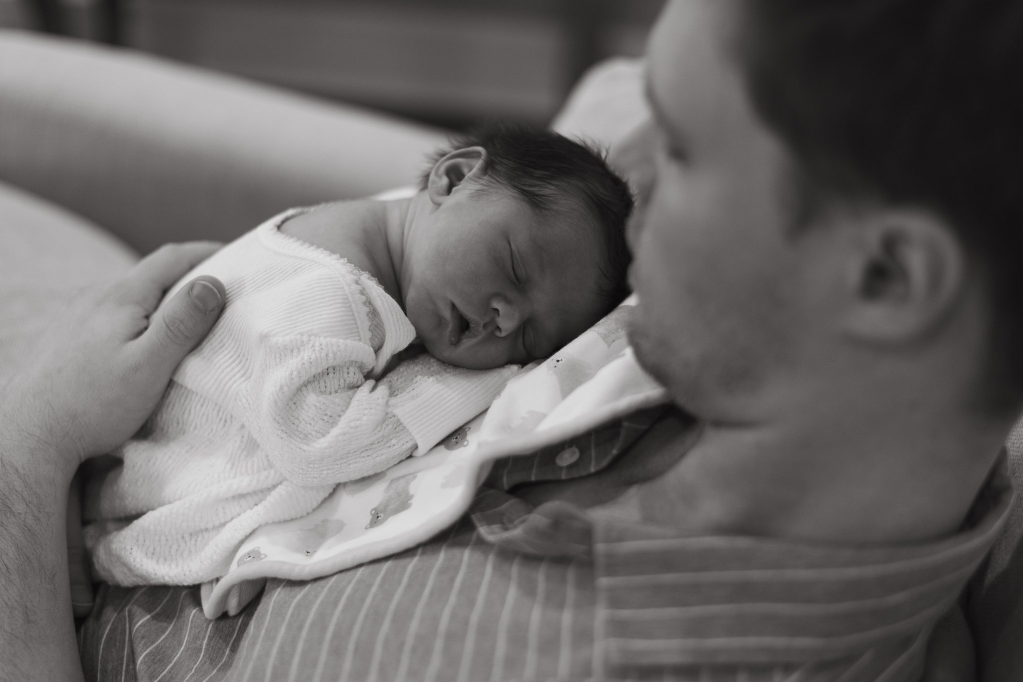 A newborn baby with dark hair sleeping peacefully on a bed, supported by an adult's hand, in black and white.
