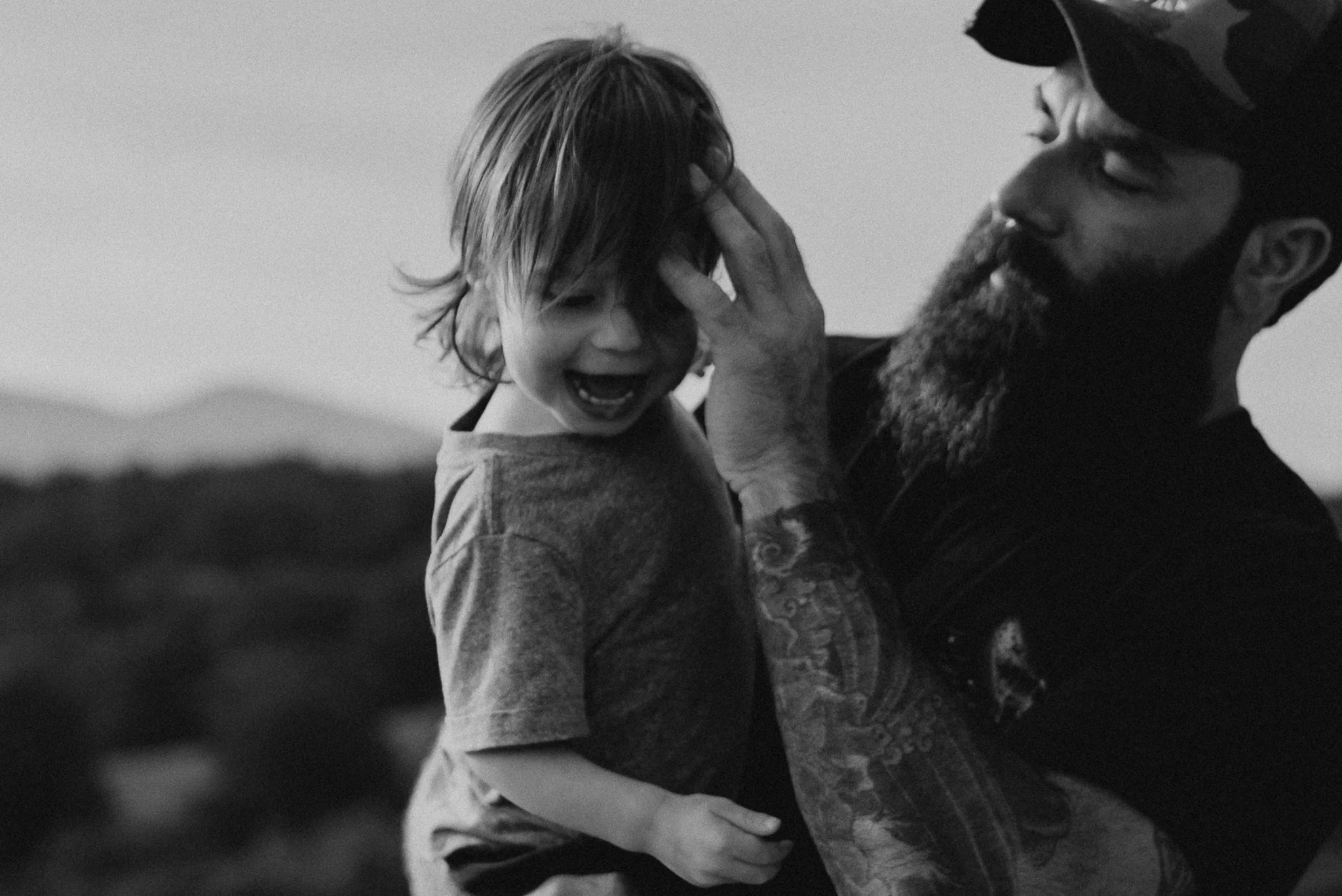 A man with a beard and a helmet is holding a young child with wet hair, and they appear to be smiling or laughing together outdoors.