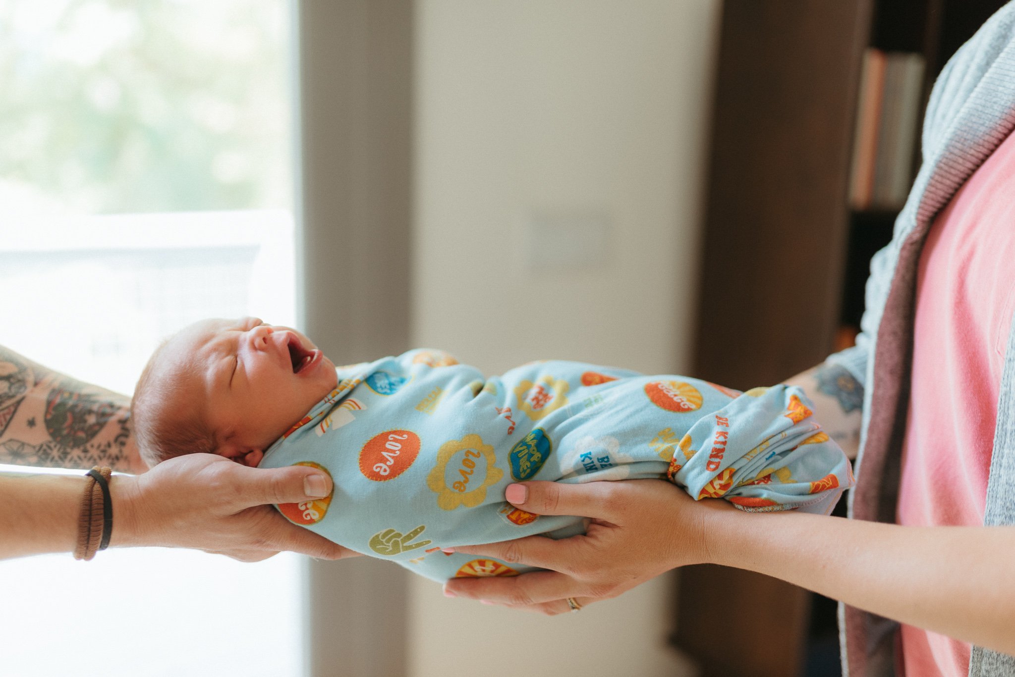 A newborn baby being gently held by an adult's hands, lying in a person's arms, wearing colorful clothing with text and patterns, with a blurred indoor background.