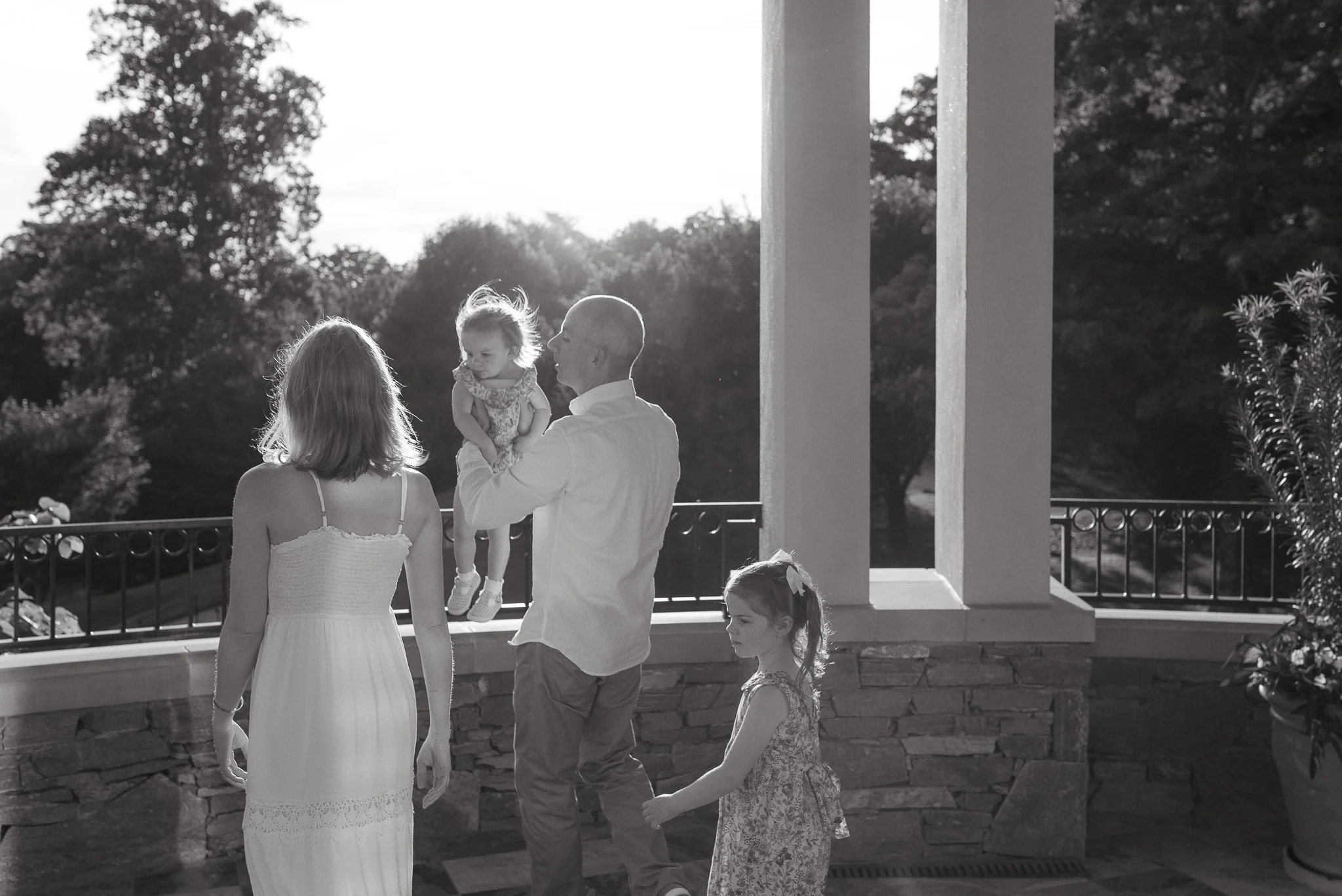 A man and a woman standing outside on a balcony with trees in the background. The man is holding a young girl, and there are two other young girls nearby, all wearing dresses. The scene is illuminated by sunlight.