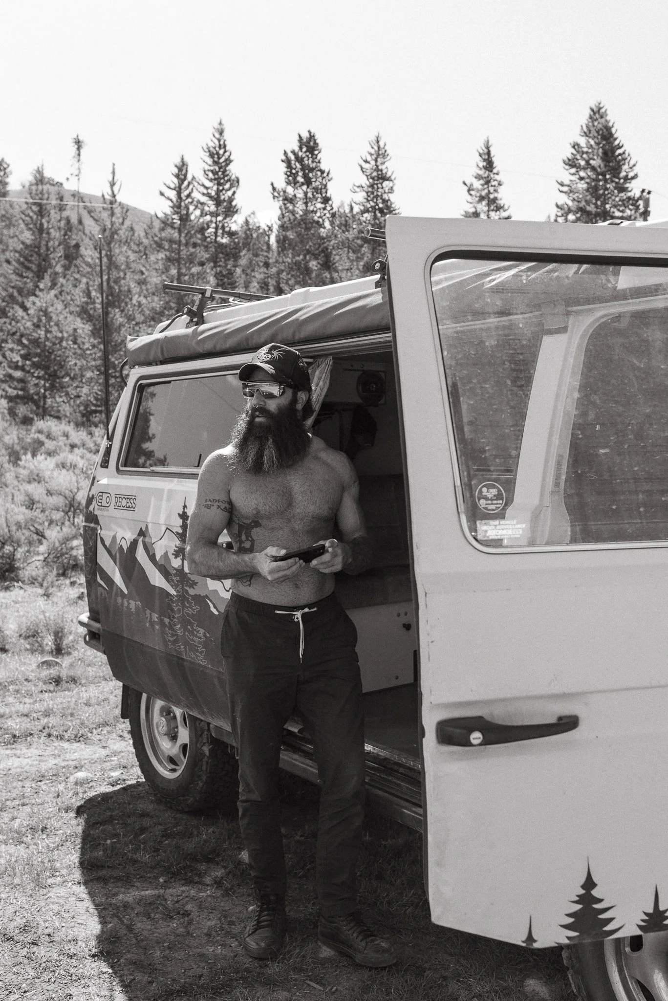 A shirtless man with a beard wearing sunglasses, a cap, and pants, standing next to a camper van with a mountain and forest background.
