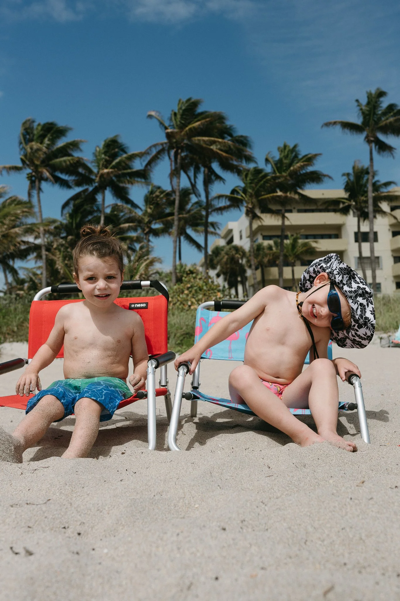 Two children in swimsuits sitting on beach chairs in the sand, with palm trees and a building in the background, under a partly cloudy sky.