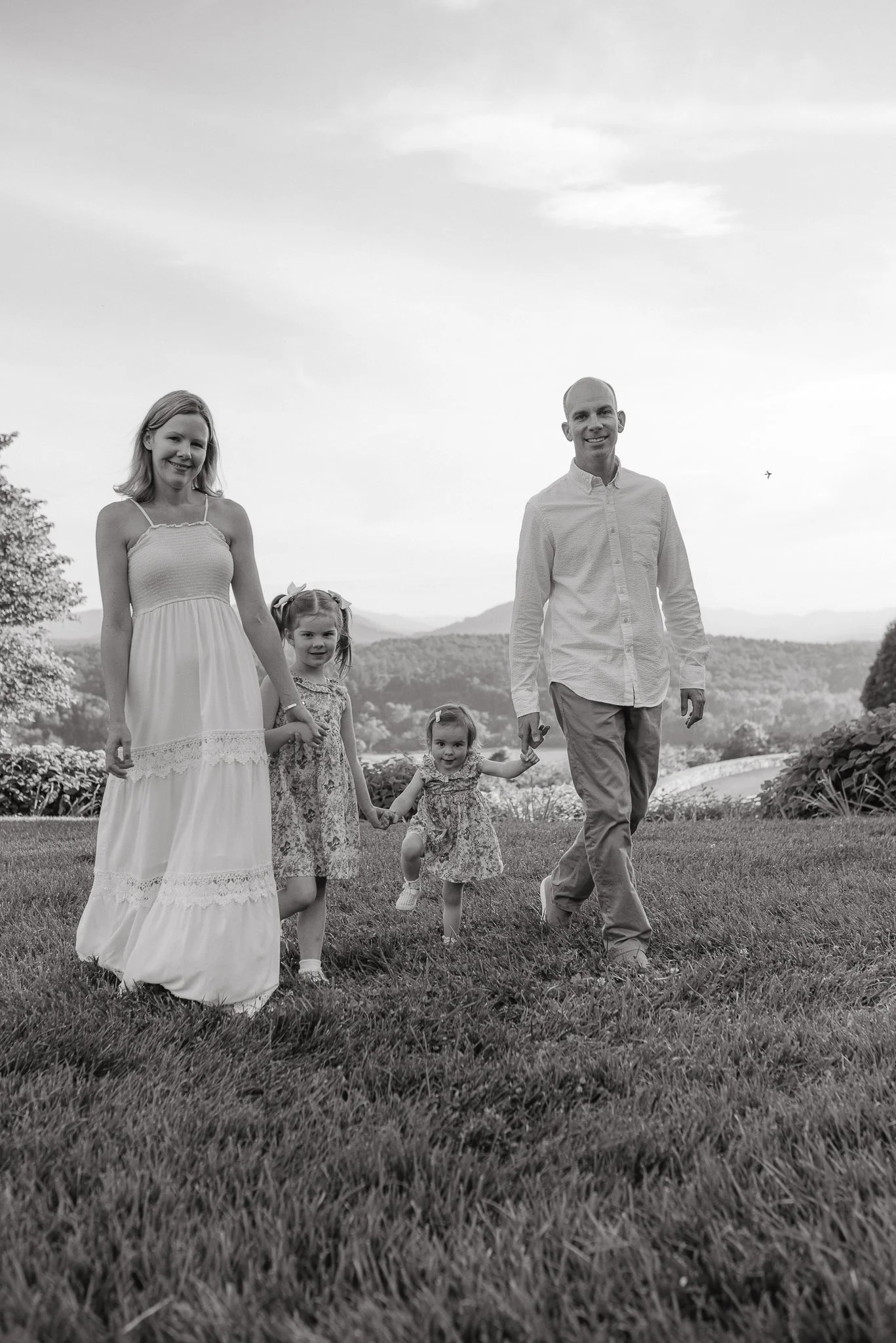 A family of five walking outdoors in a grassy area with hills and trees in the background. The family includes two adults and three children, all smiling and holding hands. The photo is in black and white.