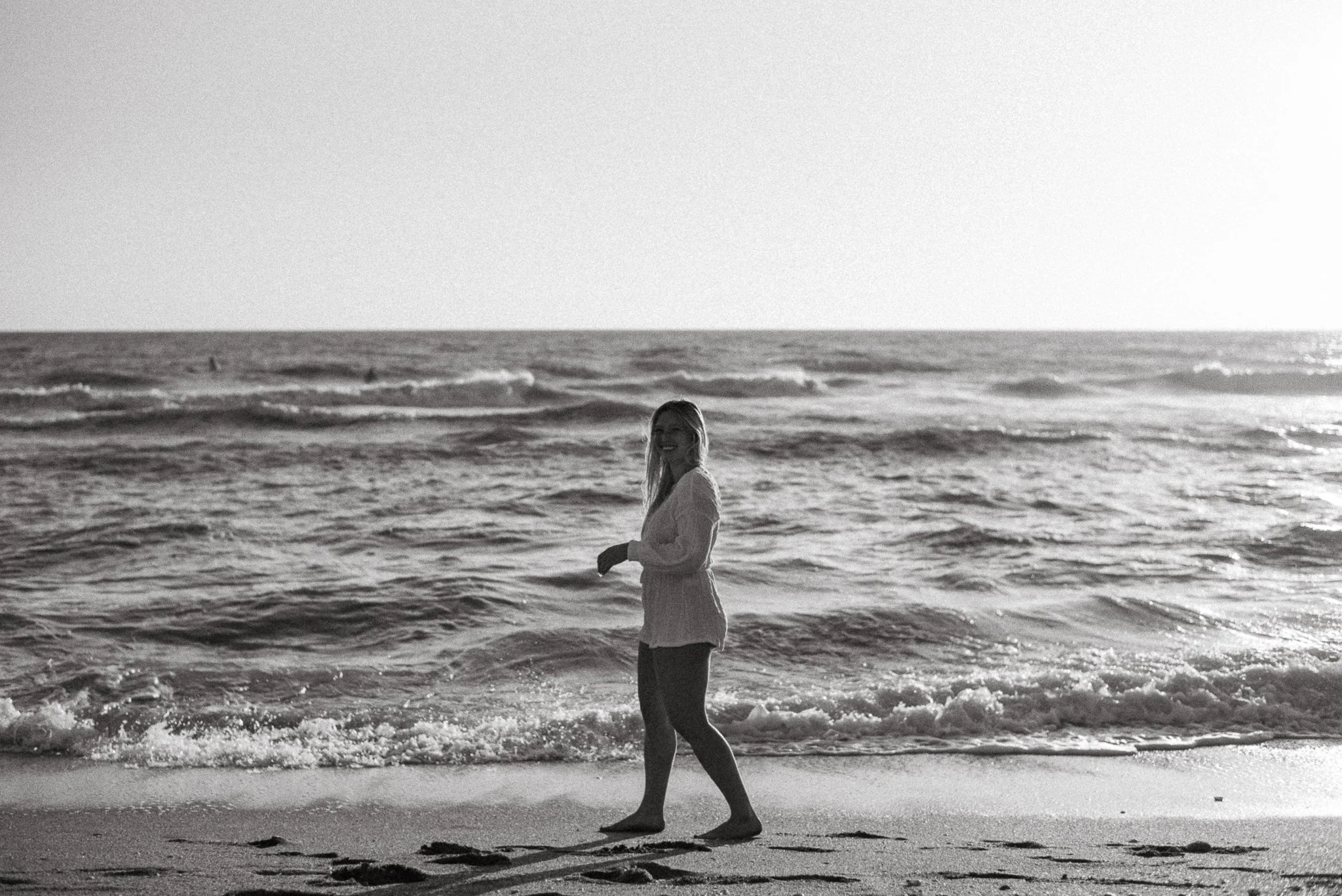 A woman walking along the beach near the ocean, smiling, with waves in the background.