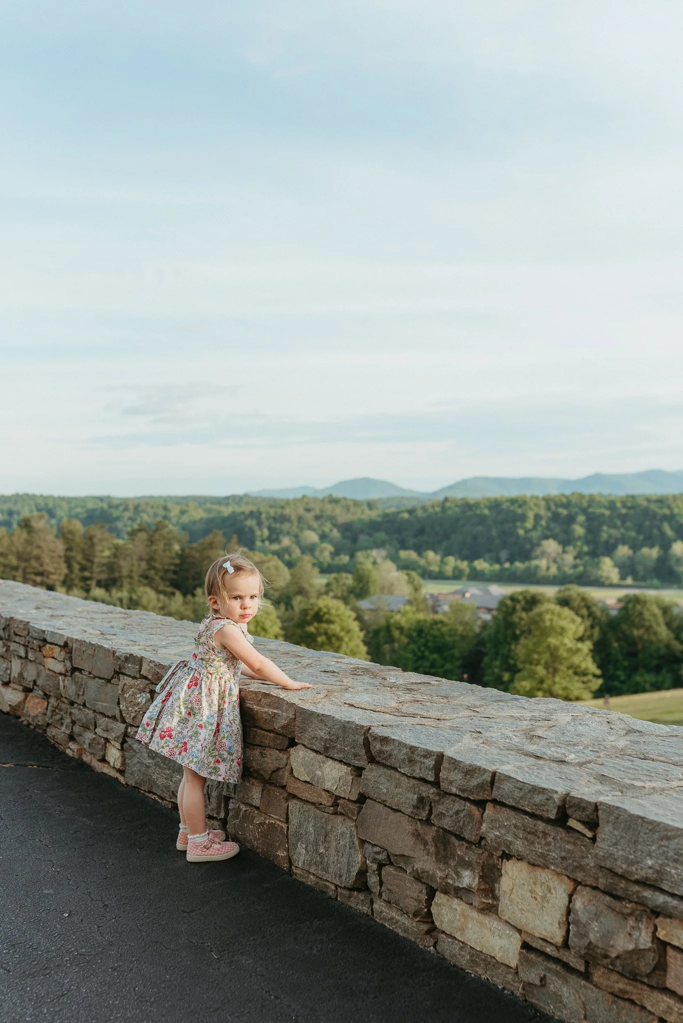A young girl wearing a floral dress and pink sneakers is standing beside a stone wall, looking over a scenic landscape of trees and rolling hills under a cloudy sky during daytime.