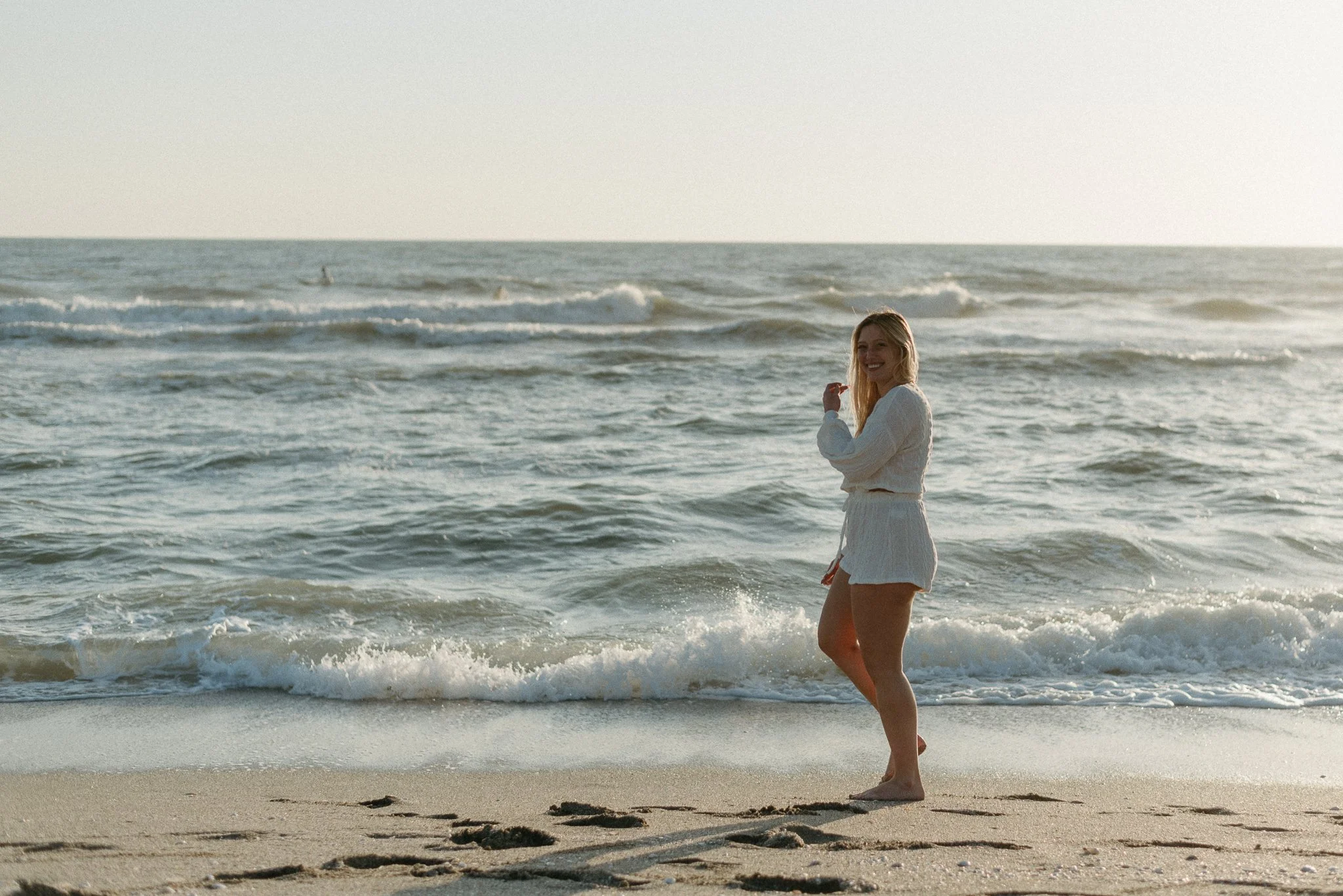 A woman in a white dress standing in the shallow water at the beach, smiling, with the ocean waves behind her and the sunset in the sky.