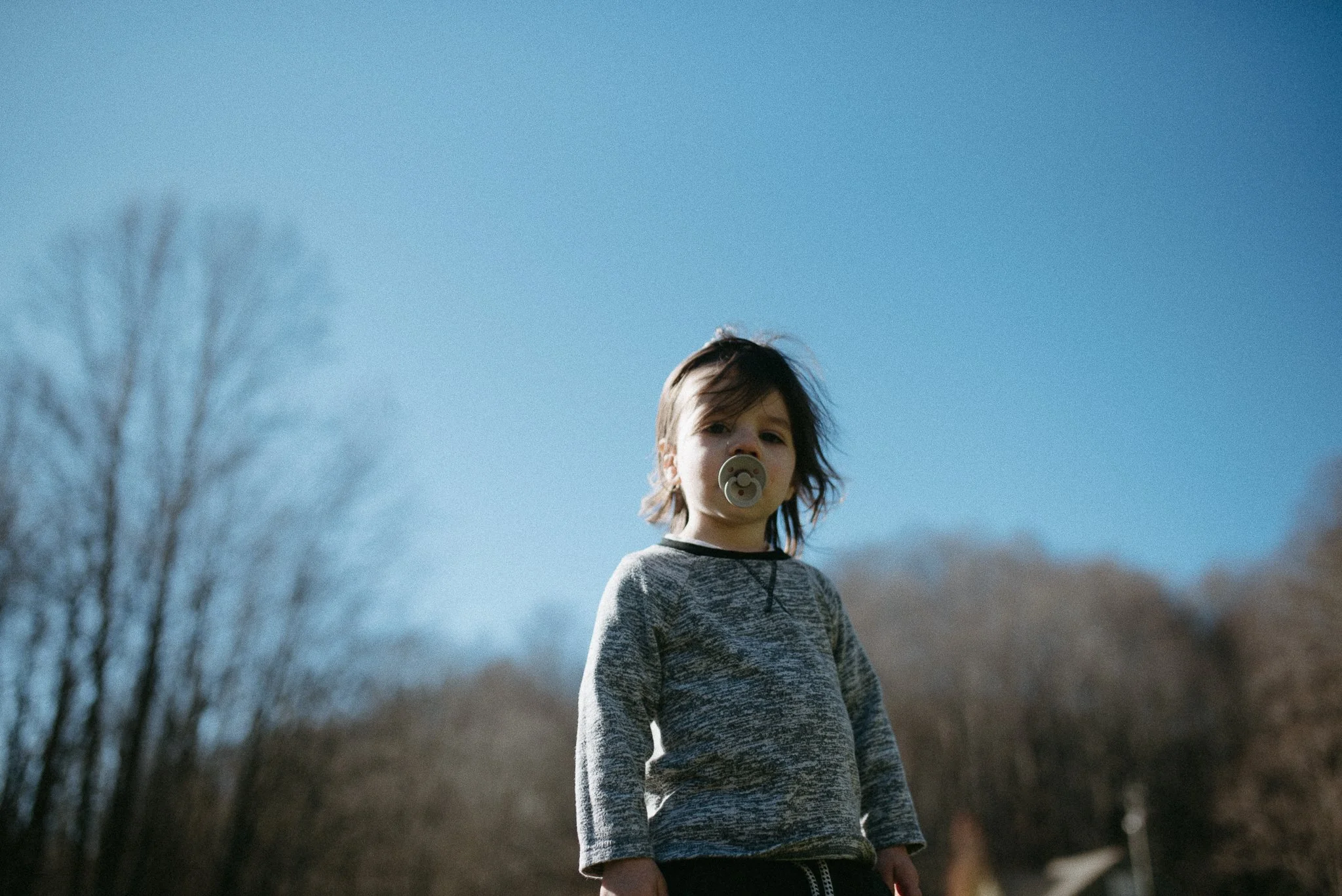 A young boy with a pacifier in his mouth standing outdoors on a clear day, with leafless trees and a blue sky in the background.