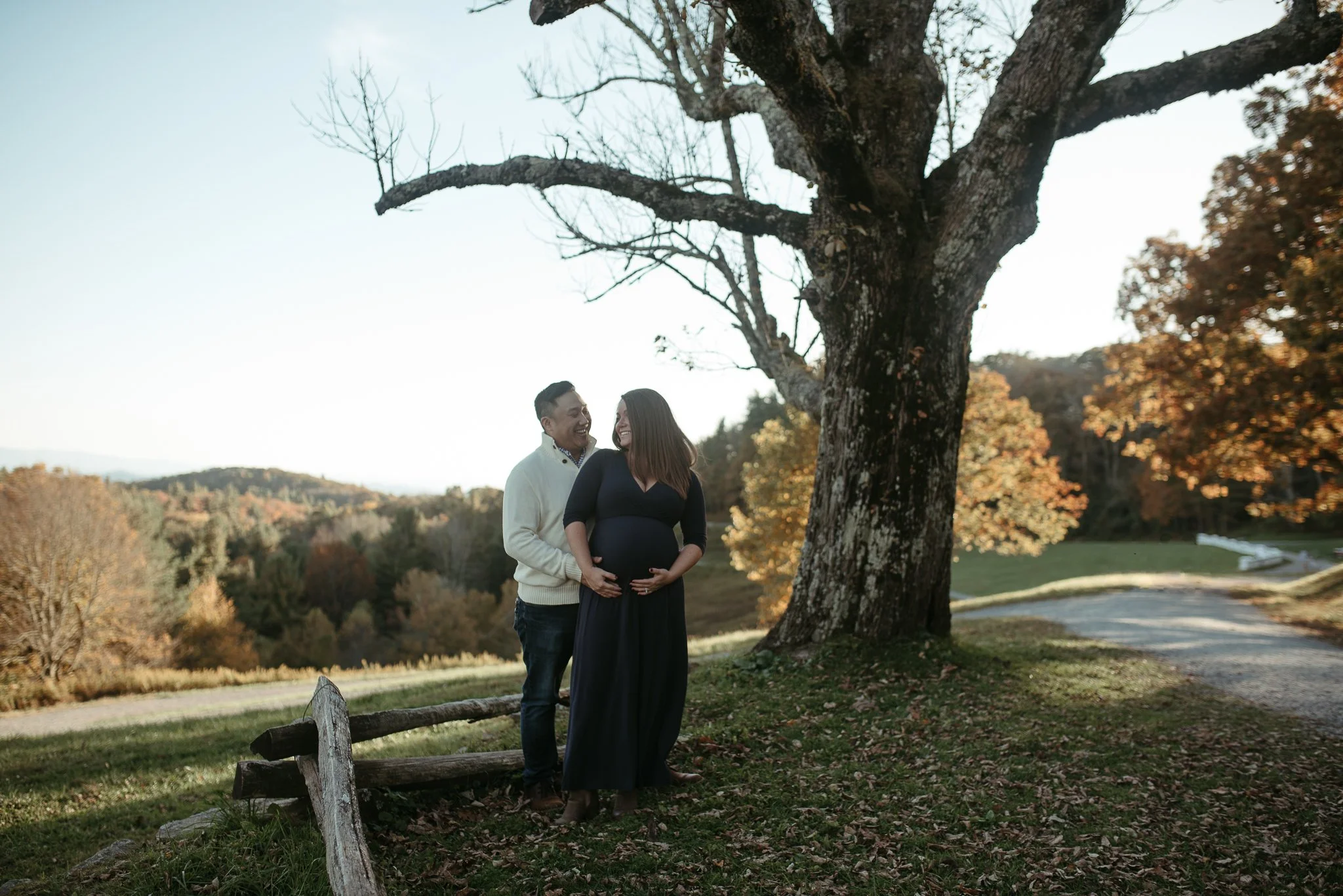 A pregnant woman wearing a long dark dress and her partner, both smiling and looking at each other, standing under a large tree in a park with autumn foliage.