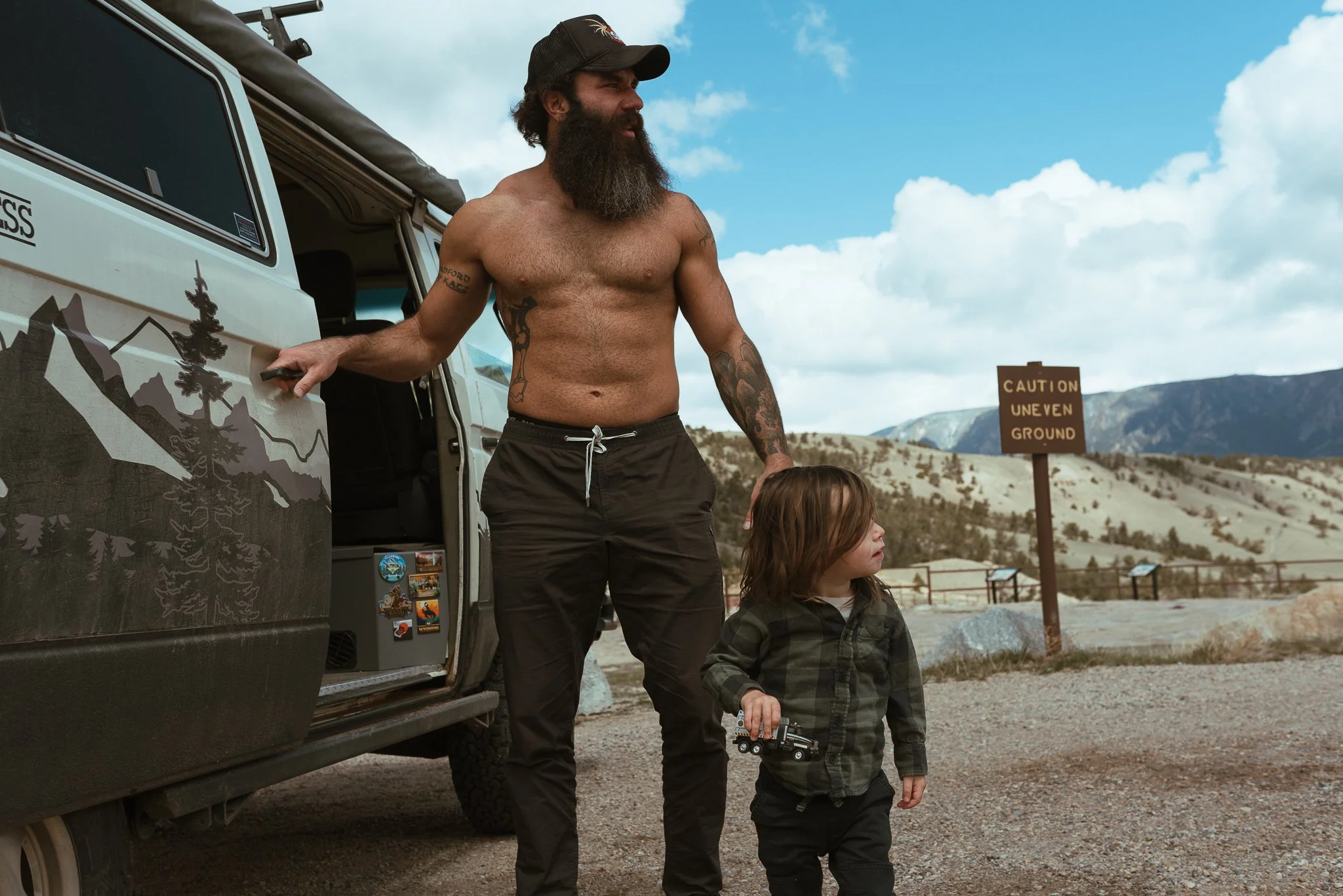 A bearded man shirtless, wearing a baseball cap, standing next to a young boy with long hair and a jacket, outdoors in a scenic area with mountains and a sign that reads 'Caution Uneven Ground'.