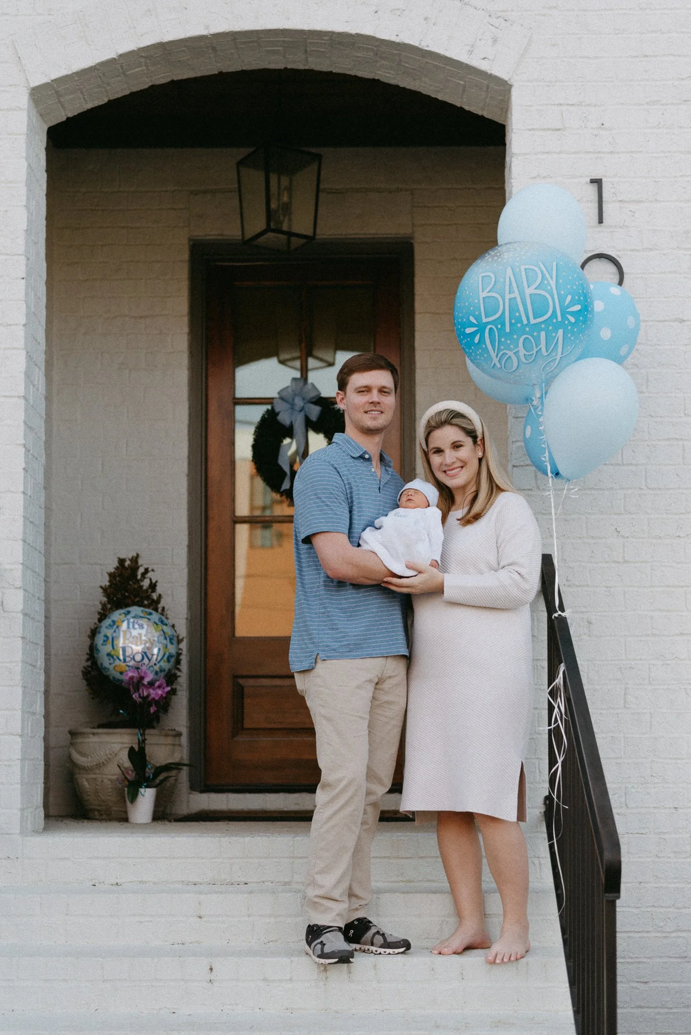 A couple standing on the front porch of their home, holding a newborn baby. The man is wearing a blue striped shirt and khakis, while the woman is in a white dress with a headband. There are blue balloons with 'Baby Boy' written on them on the right 