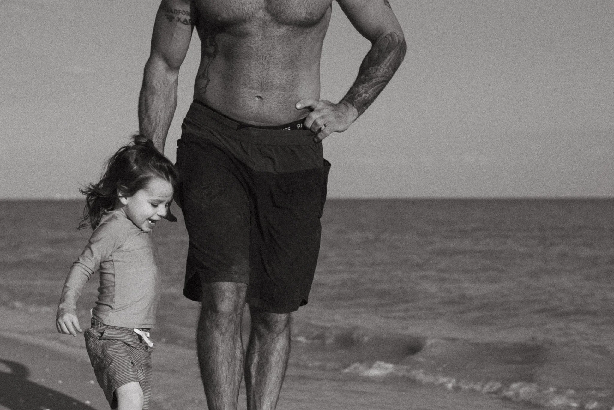 A shirtless man walking on the beach with a young girl, holding hands and smiling.