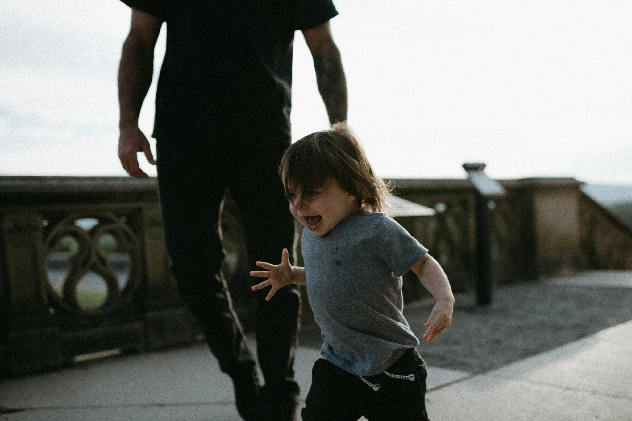 A young boy with brown hair and gray shirt laughing and playing on a rooftop with an adult wearing black clothing, with a gray sky and rooftop railing in the background.