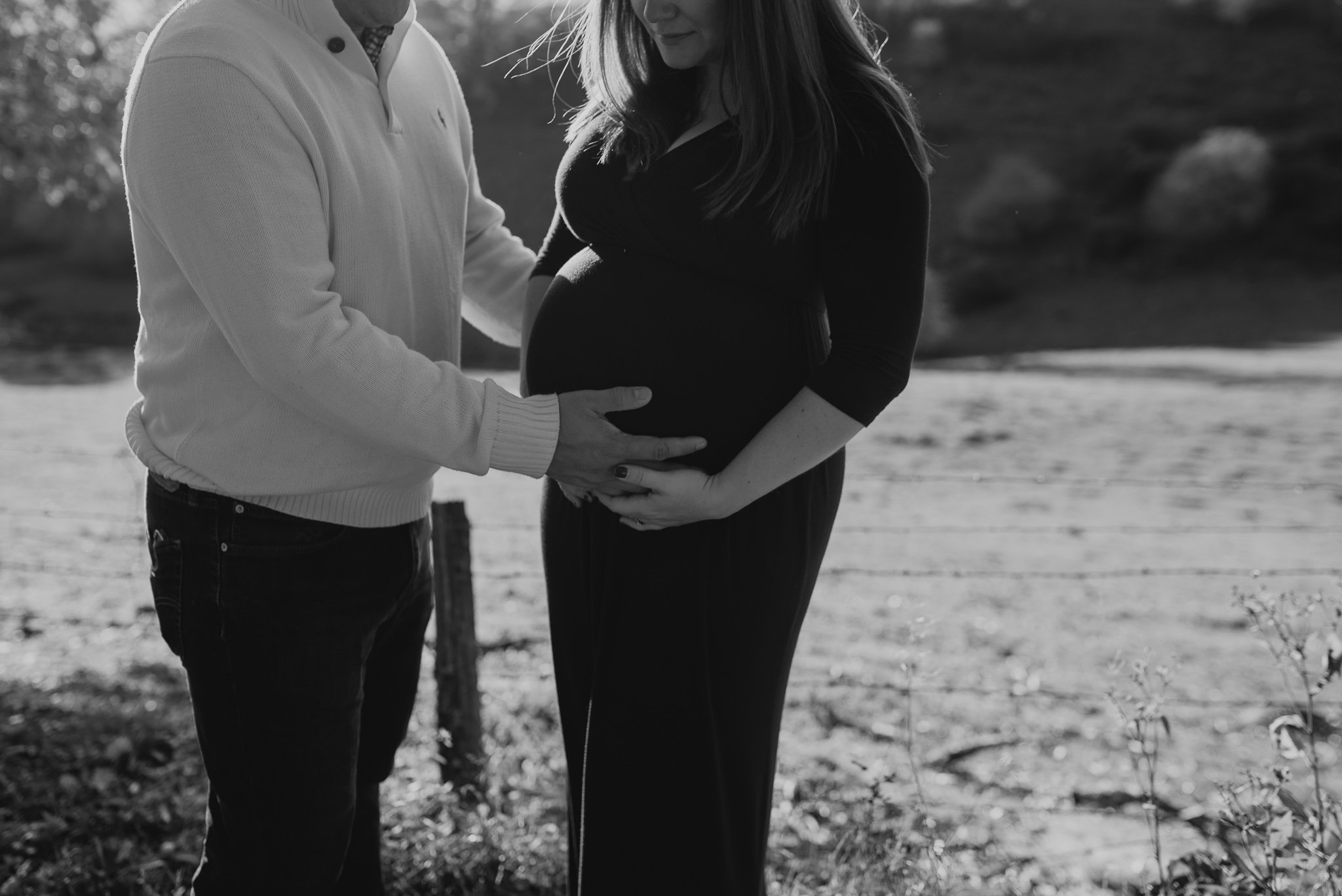 A pregnant woman in a black dress and a man in a light sweater stand close together outdoors, holding and touching her belly as they look at each other.