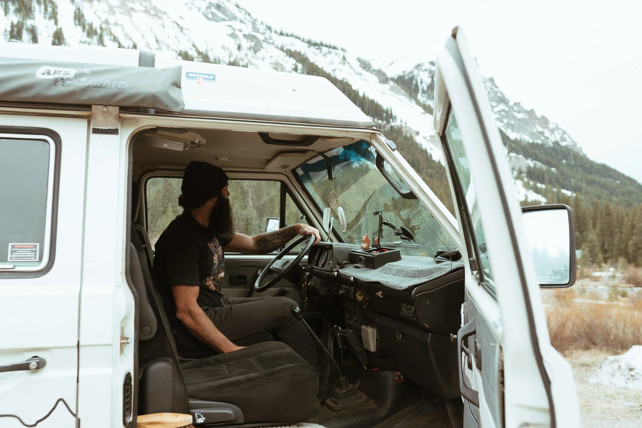 A man with a beard and black beanie sitting in the driver's seat of a white camper van, with mountains and snow in the background.
