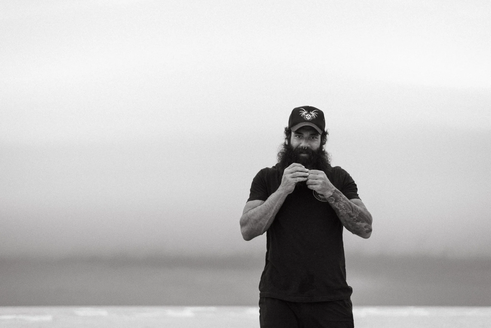 A man with a beard and tattoos on his arm, wearing a black shirt and a black cap with a graphic, stands outdoors against a cloudy sky. He is smiling and holding an object close to his face.