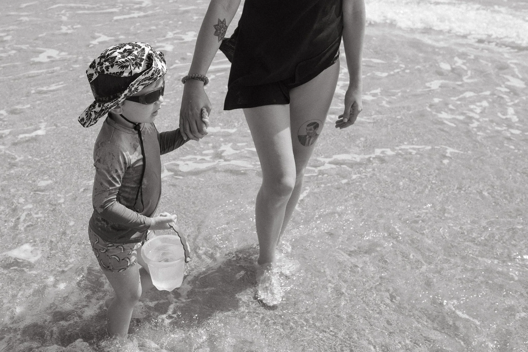 A child holding a woman's hand at the beach, with the woman wearing a black swimsuit and the child wearing a sun hat, sunglasses, and patterned swim shorts, carrying a bucket in the water.