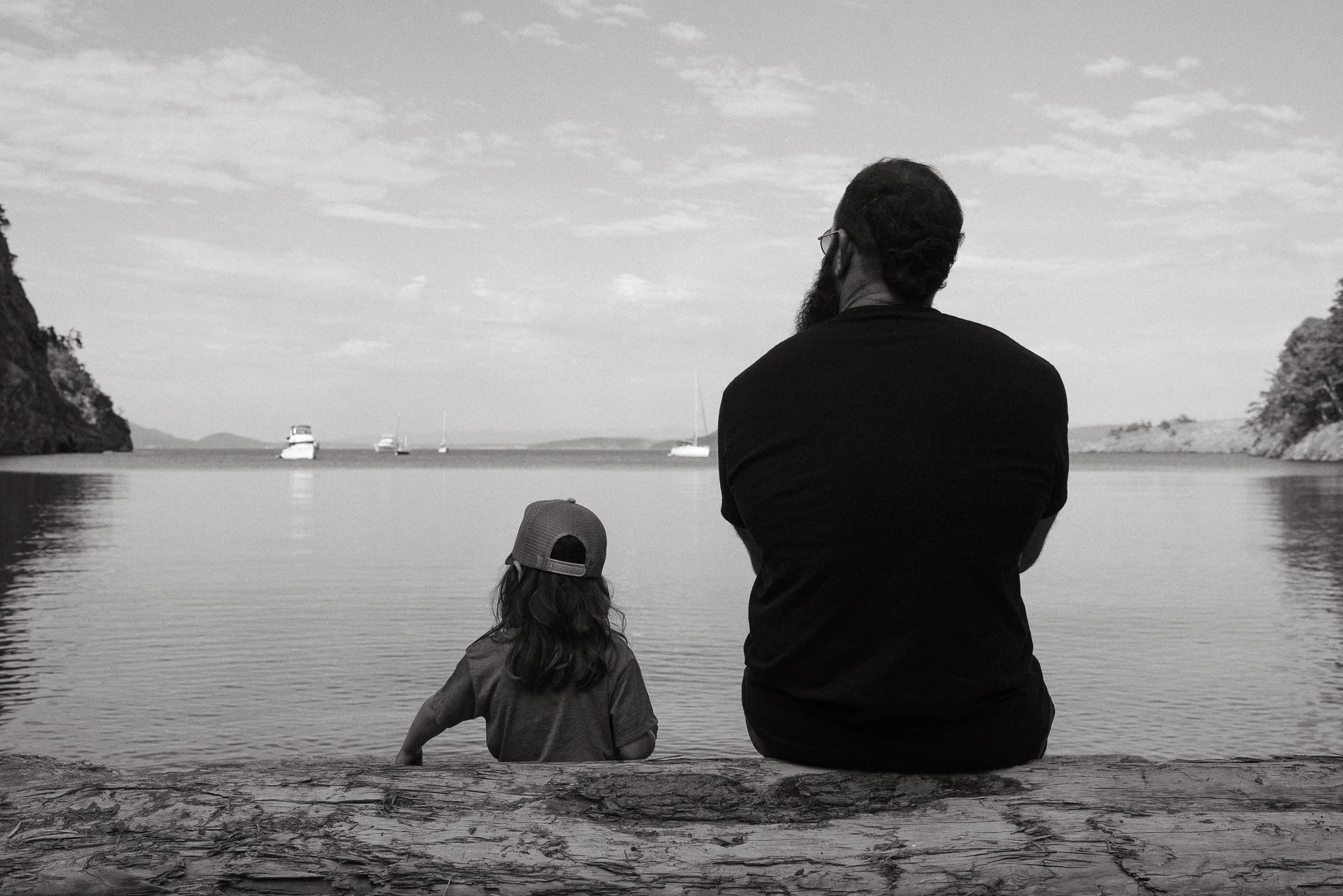 A man and a young girl sitting by the water, facing away, with boats in the distance and a rocky shoreline and trees on either side.