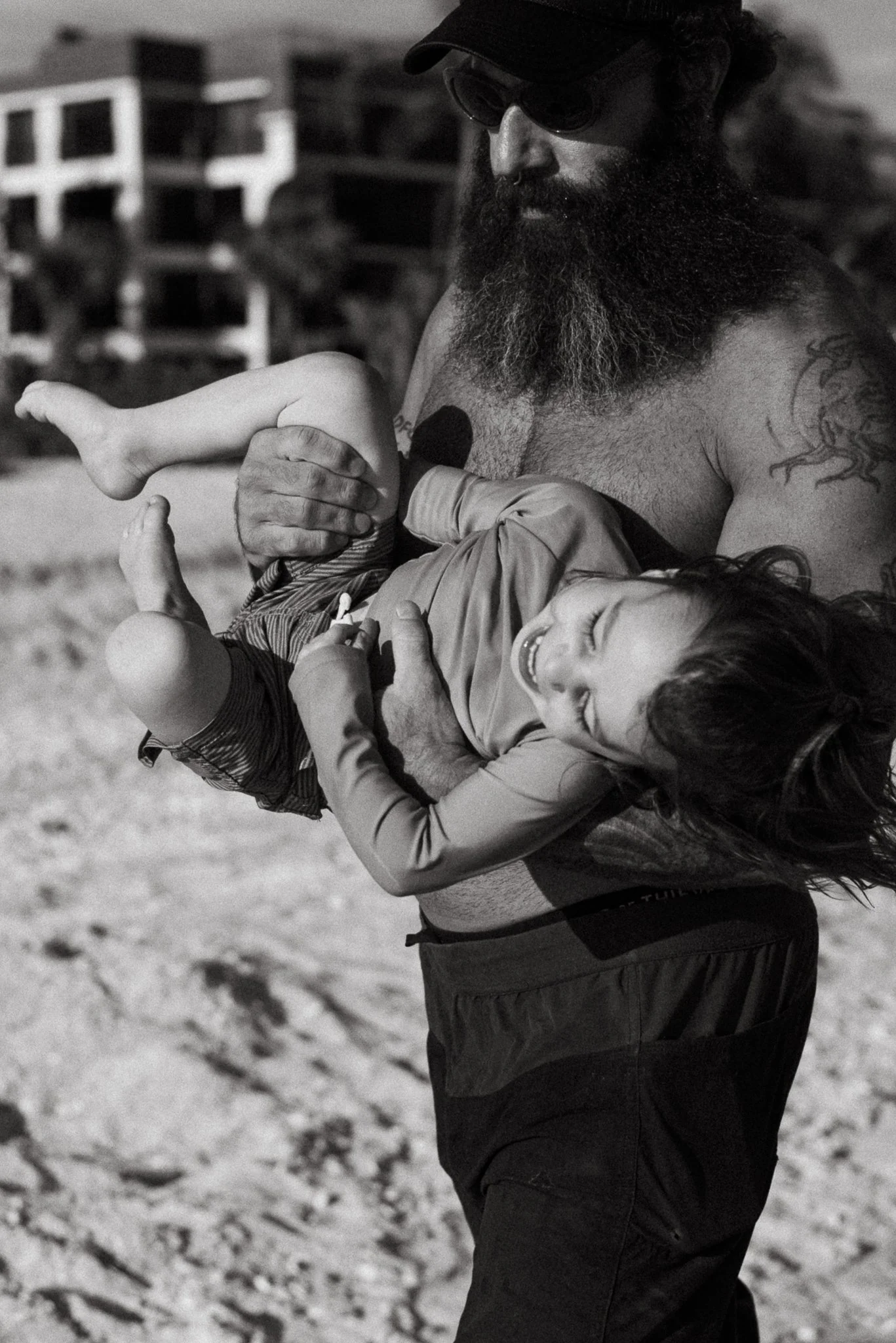 A man with a large beard, wearing sunglasses and a cap, is holding a young girl with long hair who is smiling and laughing. The girl appears to be hugging the man, playing on a sandy or gravelly surface.
