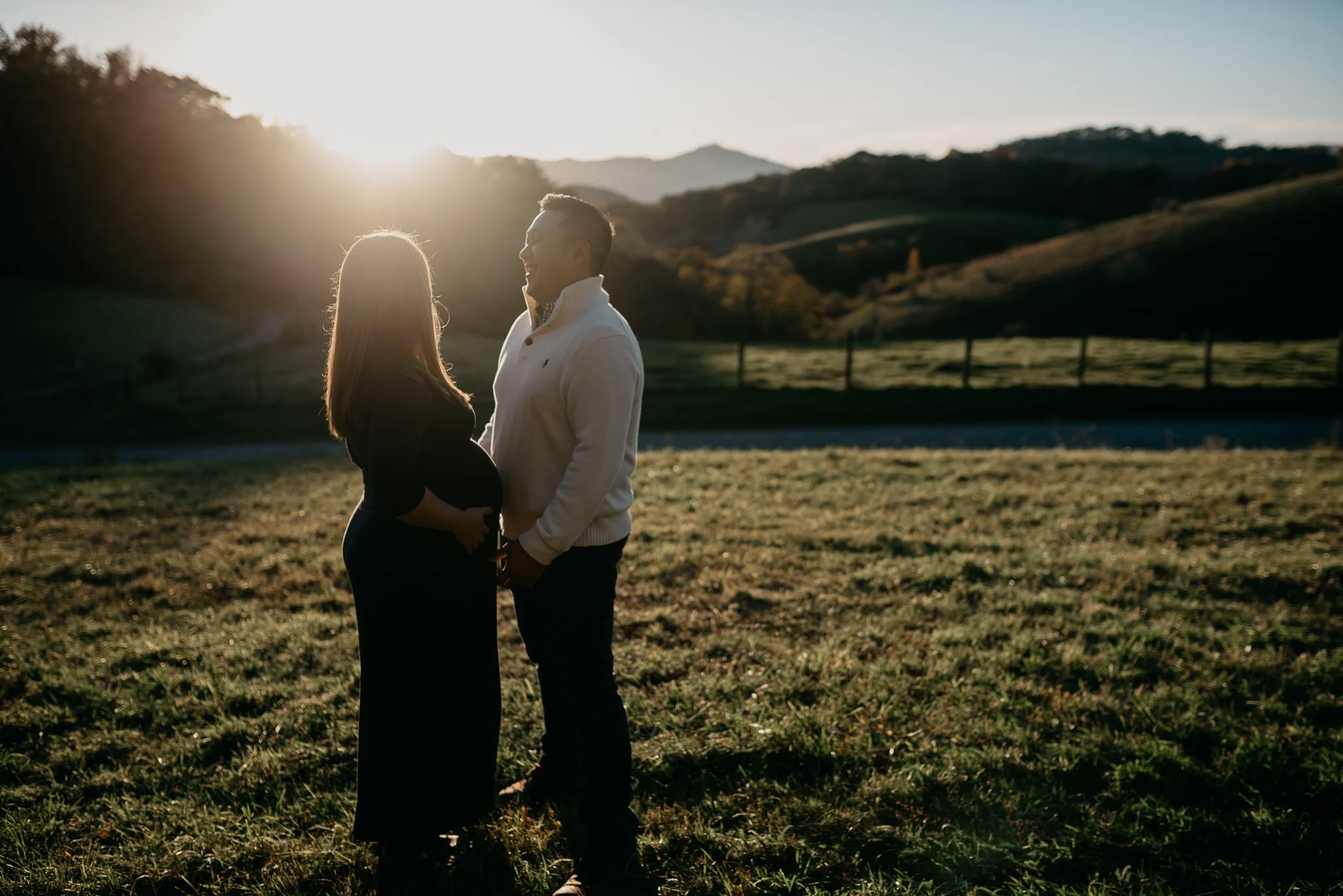 A couple standing outdoors at sunset, facing each other, the woman pregnant, with a scenic background of rolling hills and mountains.