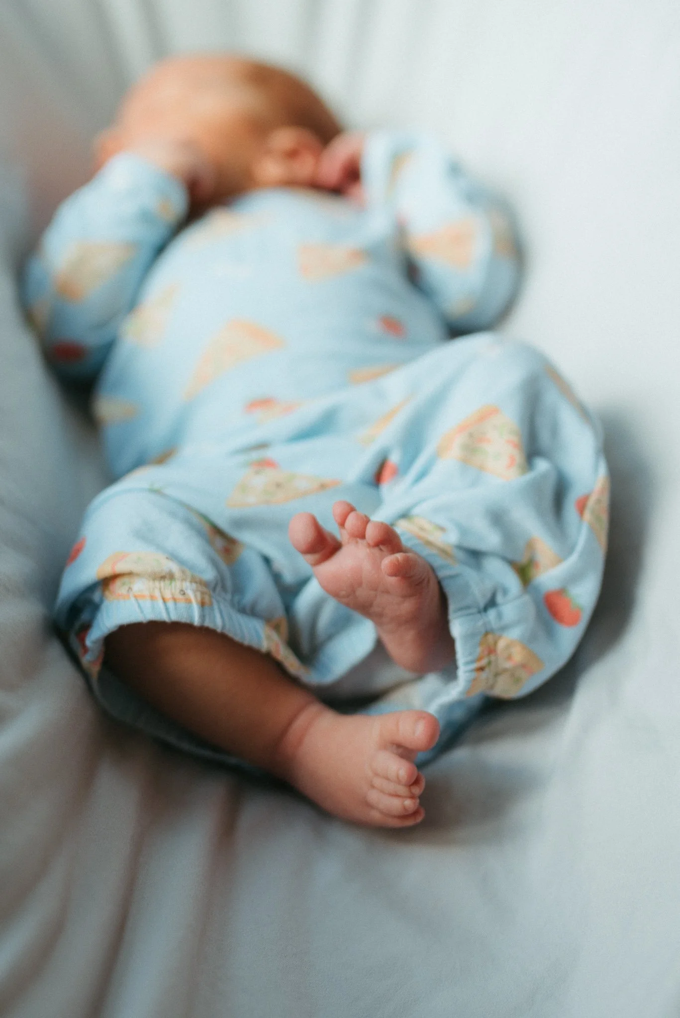 A newborn baby wearing a light blue outfit with colorful patterns, lying on a white surface, with a hand gently holding its foot.