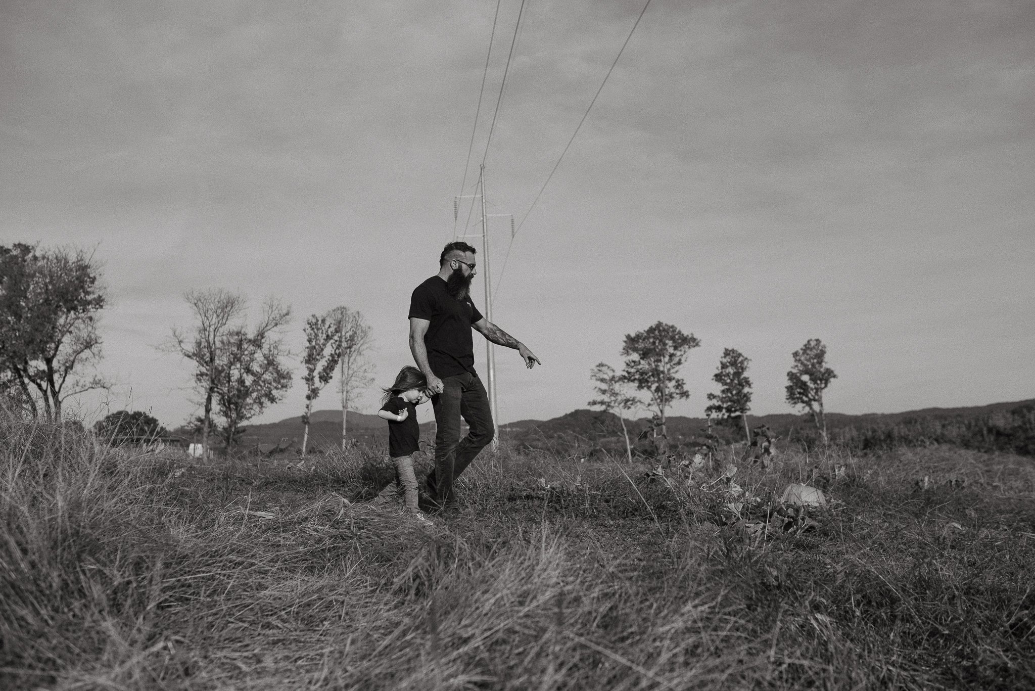 A man with a beard and glasses and a young girl are walking hand in hand through a grassy field. The man is pointing at something in the distance. The scene is in black and white, with trees and power lines in the background.