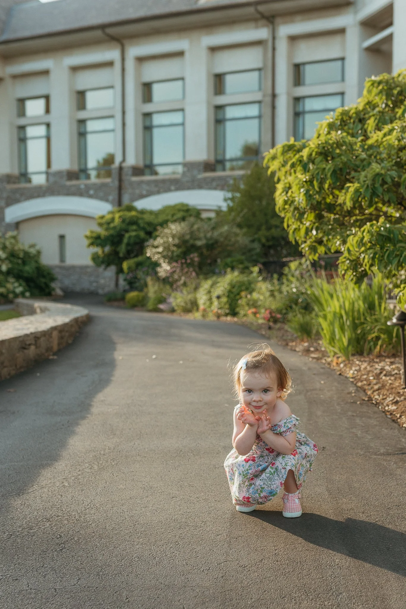 A young girl with curly hair kneeling on an asphalt driveway, wearing a floral dress and pink checkered shoes, smiling at the camera with her hands under her chin, in front of a house with large windows, plants, and shrubs.