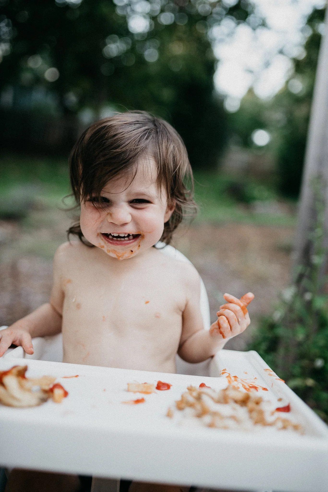 A young child with wet brown hair, smiling and holding a piece of pizza, sitting in a high chair outdoors. There is pizza and food remnants on the white tray in front of the child, with a blurred greenery background.