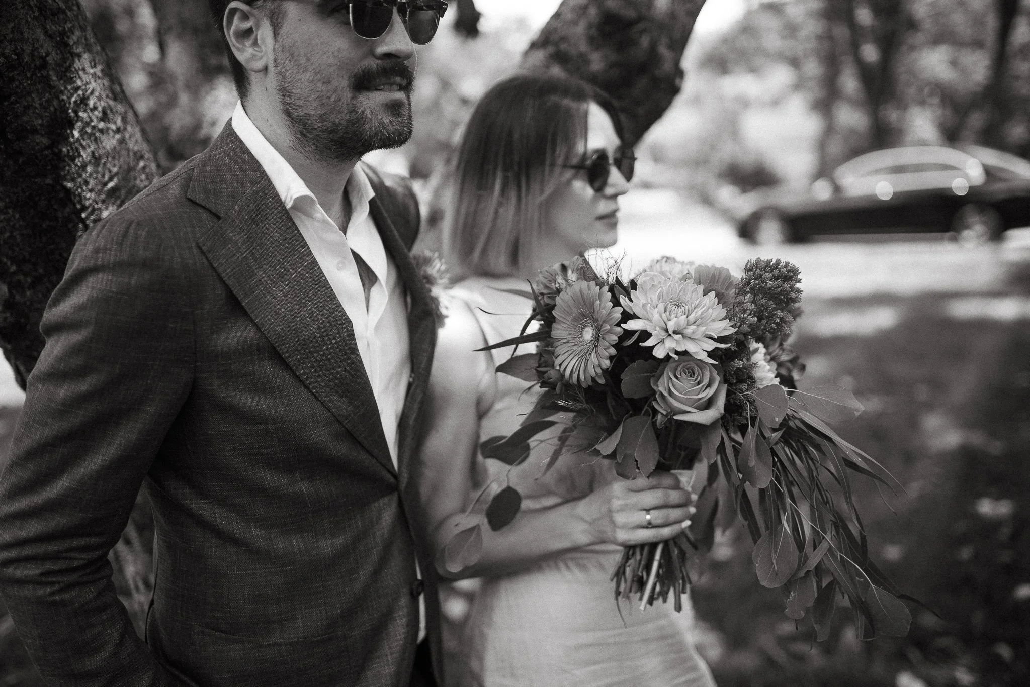 A man and woman, both wearing sunglasses, standing outdoors by trees. The woman is holding a large bouquet of flowers. The photo is in black and white.