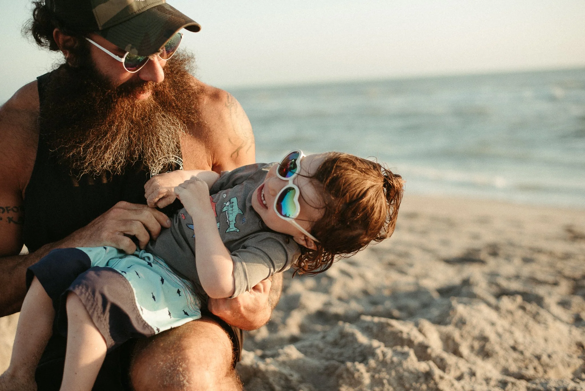 A man with a beard and sunglasses holding a smiling red-haired boy with reflective sunglasses, both sitting on the beach during sunset.