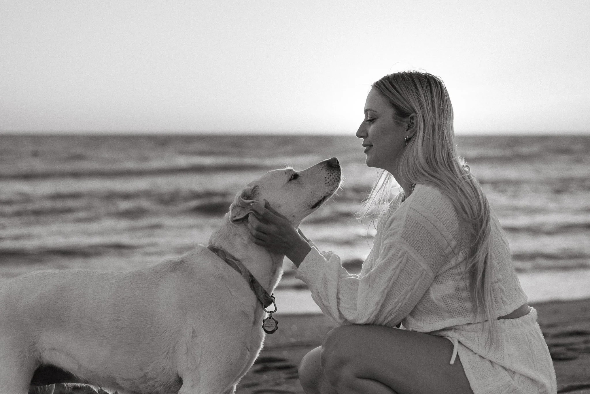 A woman with long hair wearing a light sweater kneeling at the beach, gently holding a Labrador Retriever dog by the face, both facing each other with the ocean in the background.