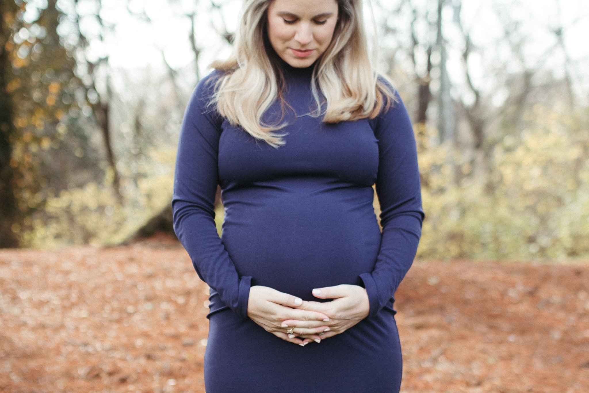 A pregnant woman in a navy blue dress and a matching jacket is standing outdoors in a forest or park with autumn leaves on the ground, holding her belly with both hands and looking down gently.