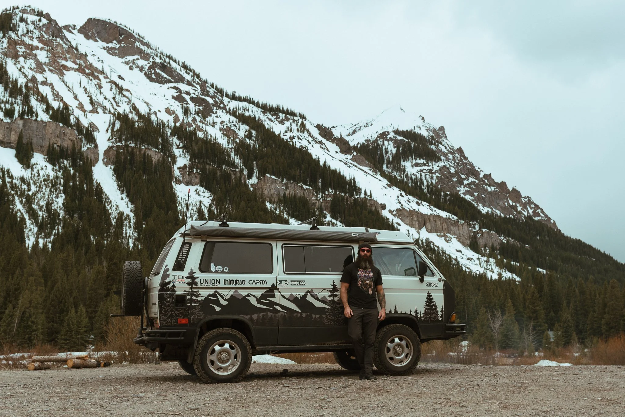 A man with a beard and glasses standing next to a black and white off-road camper van in front of snow-covered mountains with evergreen trees.