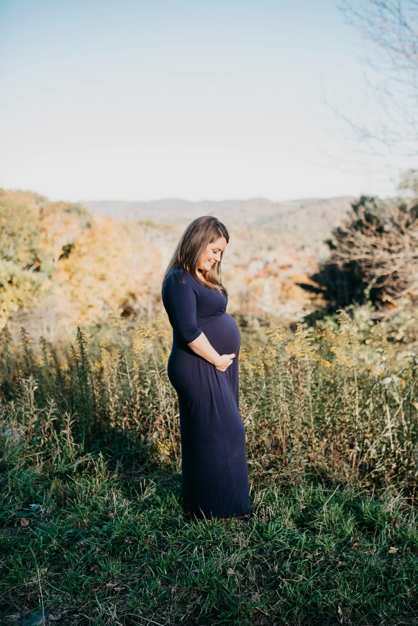 A pregnant woman in a dark blue dress is standing outdoors in a field, looking down at her belly with a gentle smile. Fall foliage and distant hills are in the background.
