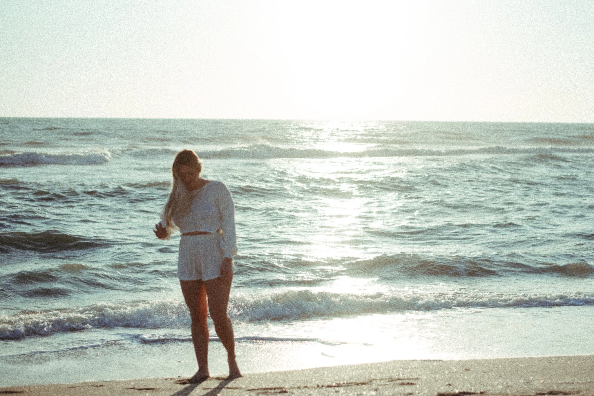 A woman in a white dress walking barefoot along the ocean shoreline at sunset, with the waves and sky in the background.