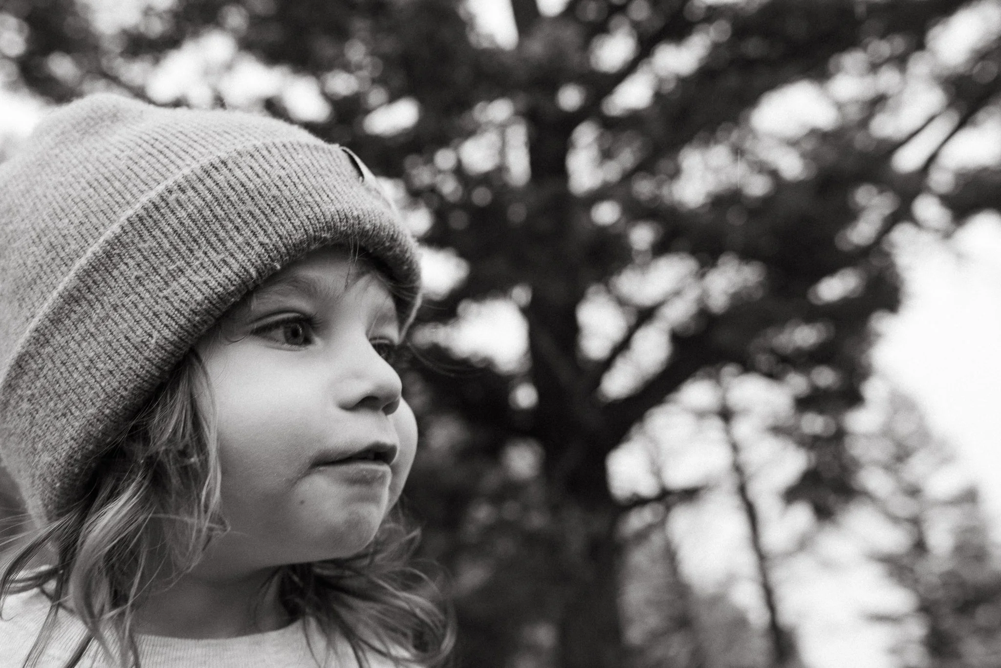 Close-up of a young girl with long curly hair wearing a knit hat, looking to the side, with a blurred background of tree branches and sky.