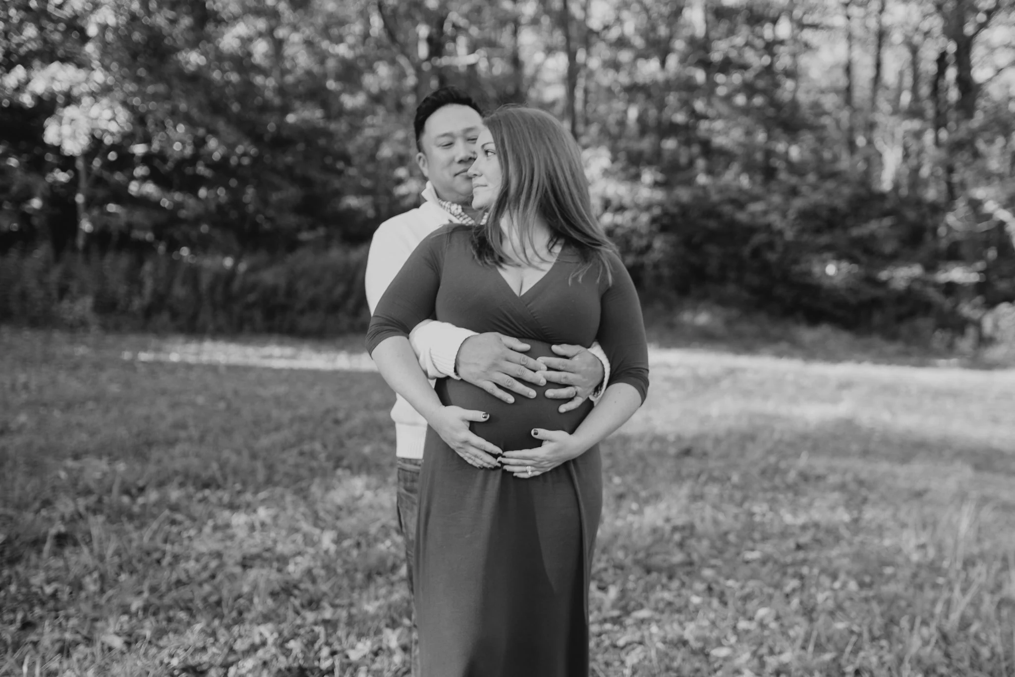 A couple, with the man embracing the pregnant woman from behind, standing outdoors in a park with trees in the background, captured in black and white.