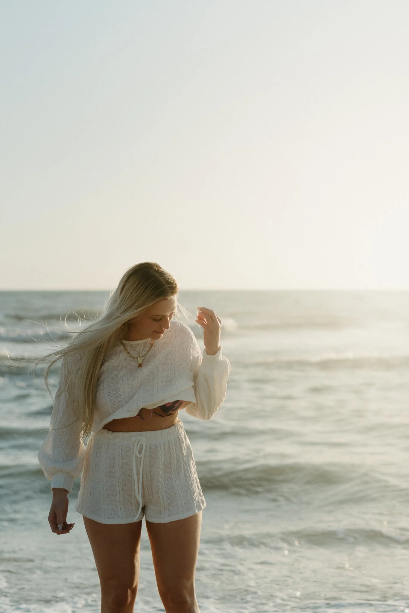 A young woman with long blonde hair standing on a beach near the ocean, looking down and touching her hair, wearing a white sweater and matching shorts in the late afternoon or early evening.