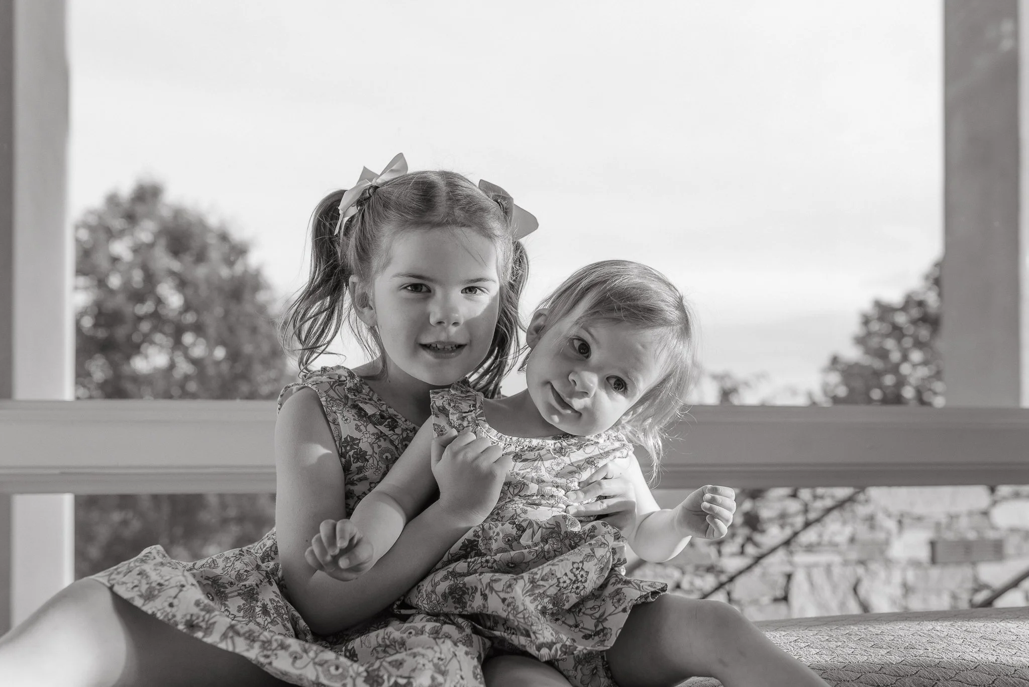 Two young girls, one with pigtails and bows, and the other with loose hair, sitting closely together outdoors and looking at the camera, with trees in the background.