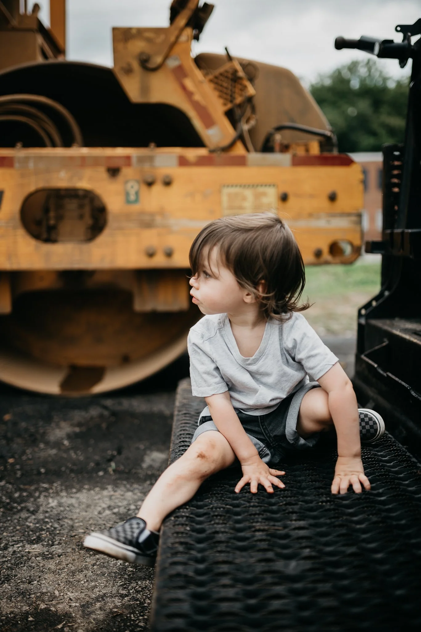 Young boy sitting outdoors on a rubber mat, dirty legs, wearing a gray t-shirt, shorts, and checkered slip-on shoes, with a large industrial toy vehicle, possibly a bulldozer or tractor, in the background.