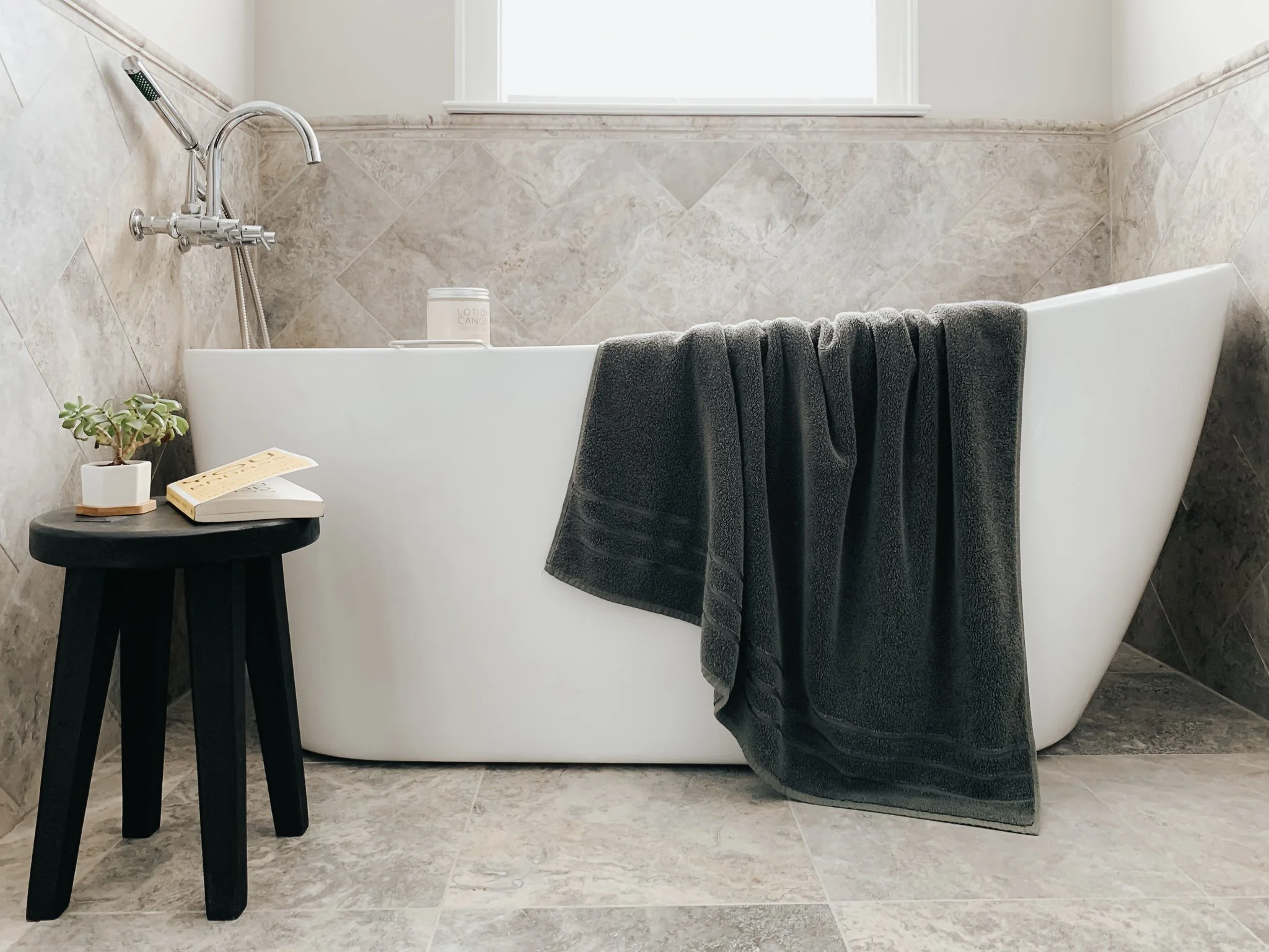 A white bathtub with a black towel draped over its side, a small black bedside table with a potted plant and a book, and a window above the tub letting in natural light. The walls and floor are tiled in beige tones.