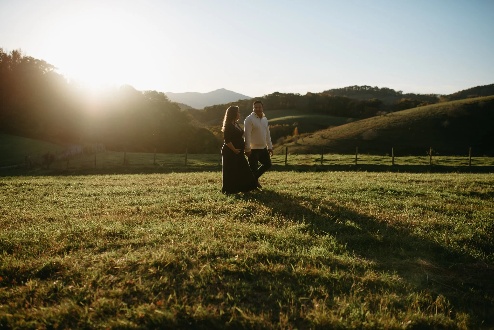 A man and woman holding hands and walking in a grassy field during sunset, with rolling hills and trees in the background.